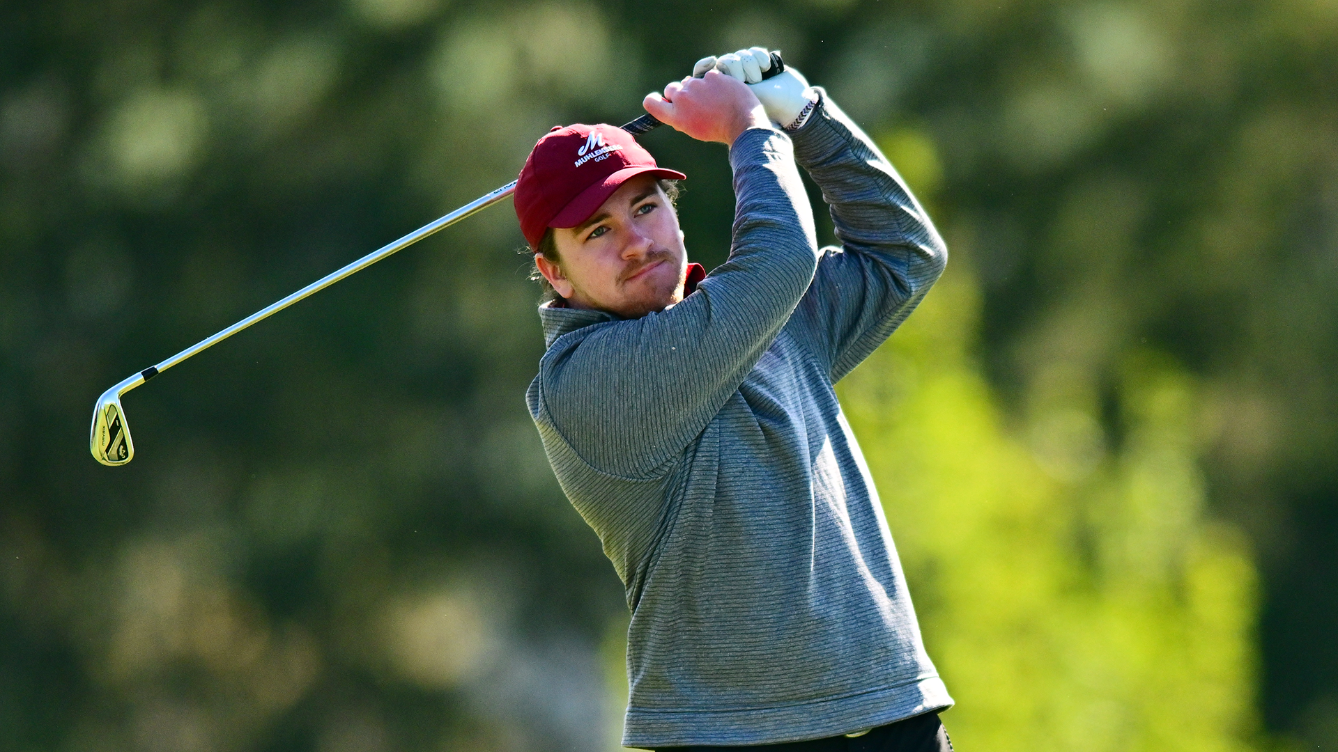a golfer in a grey longsleeve shirt and red cap follows through on his swing