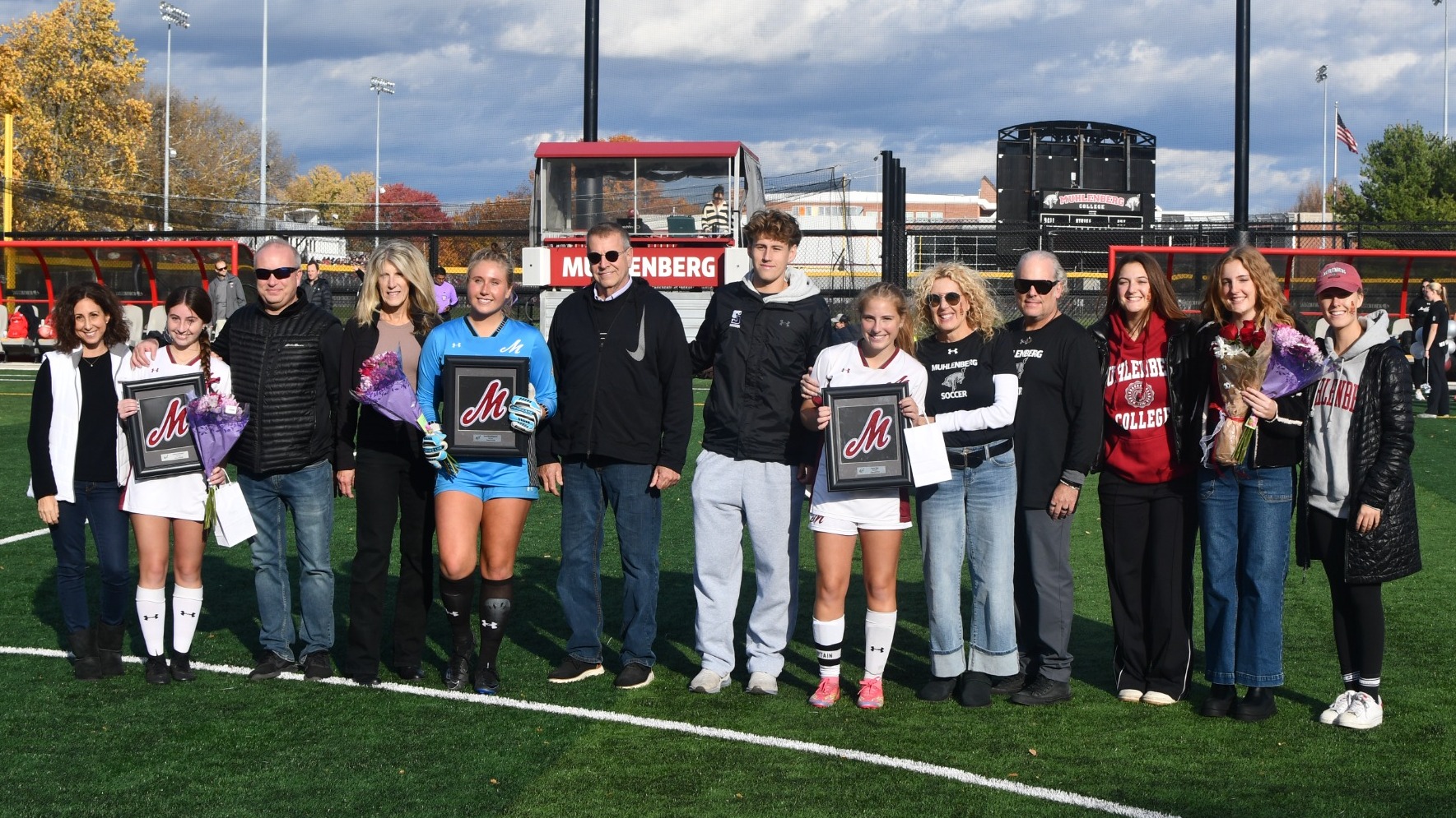 Three soccer players in white uniforms pose with their families on a soccer field 