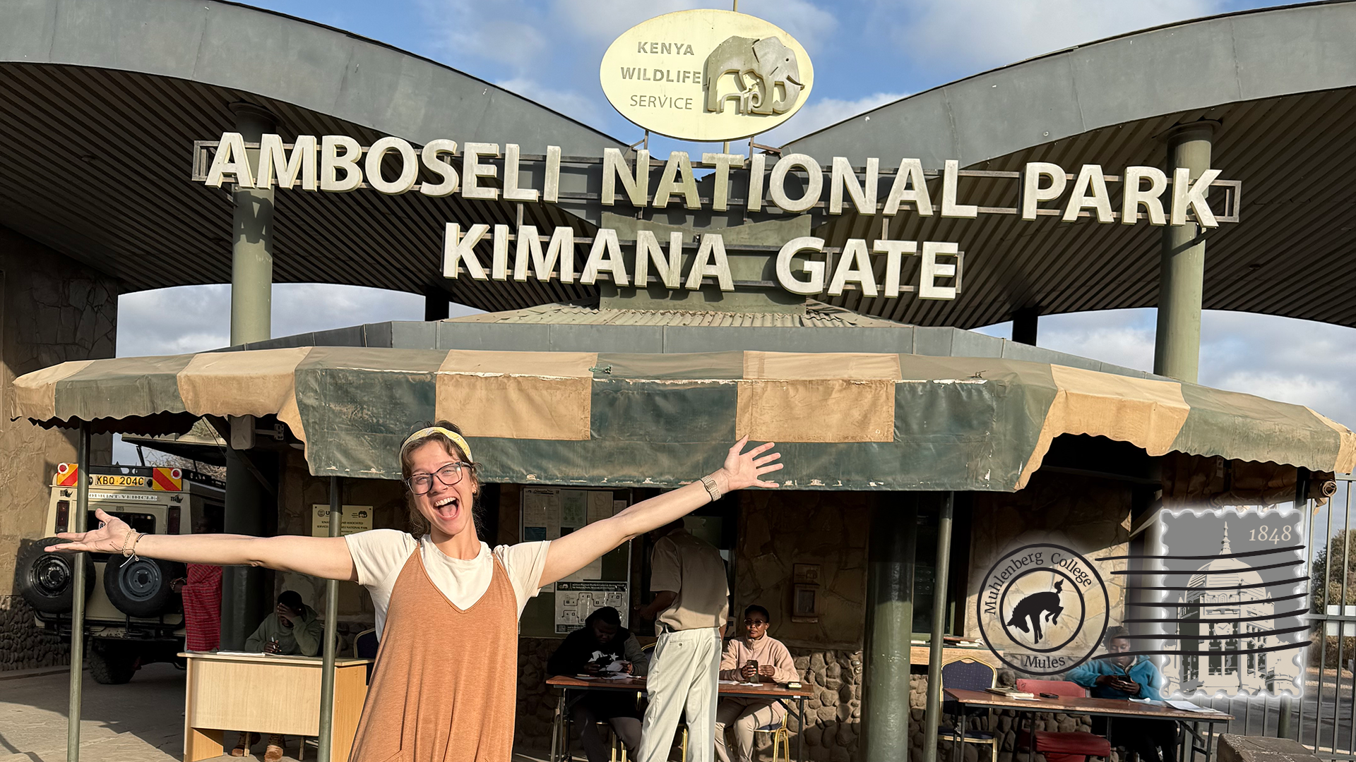 A young woman poses in front of a sign that reads Amboseli National Park Kimani Gate