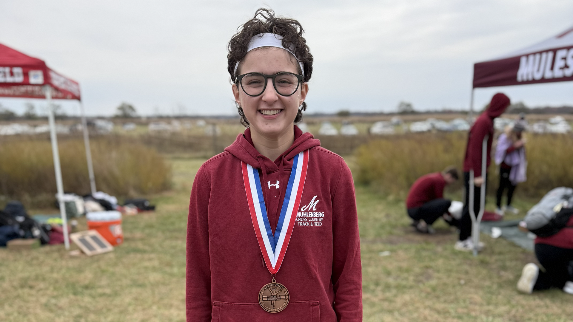 A cross country runner in a red jacket poses with a medal around their neck