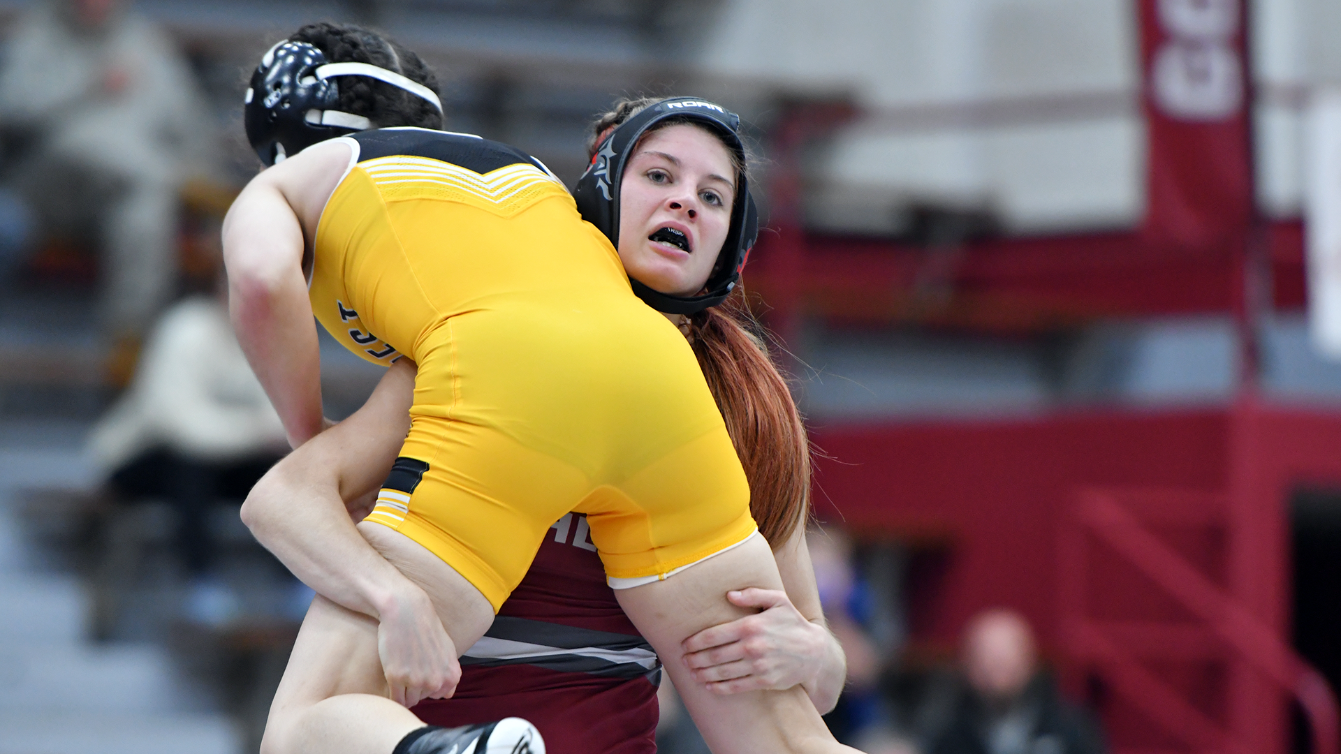 A wrestler in a red singlet lifts a wrestler in a yellow singlet off the ground