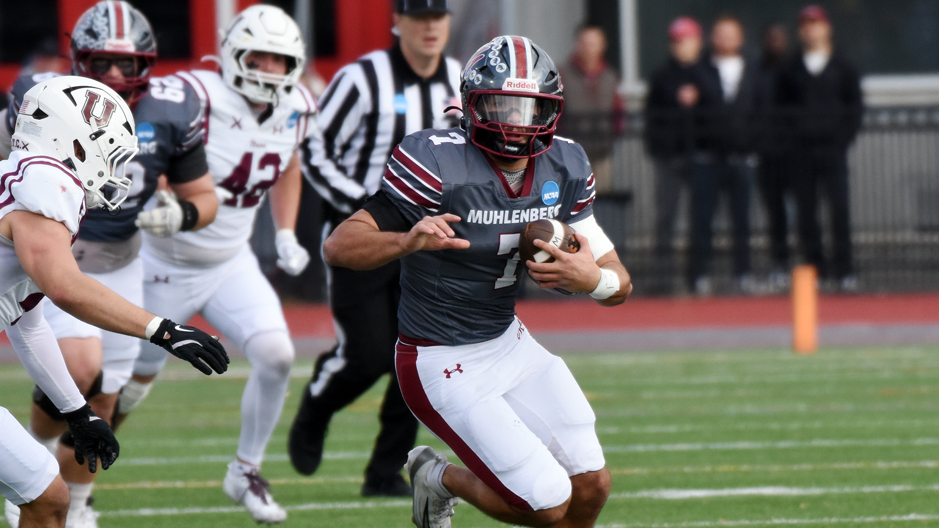 A football player in a grey jersey and silver helmet runs while being pursued by an opponent in a white jersey and white helmet