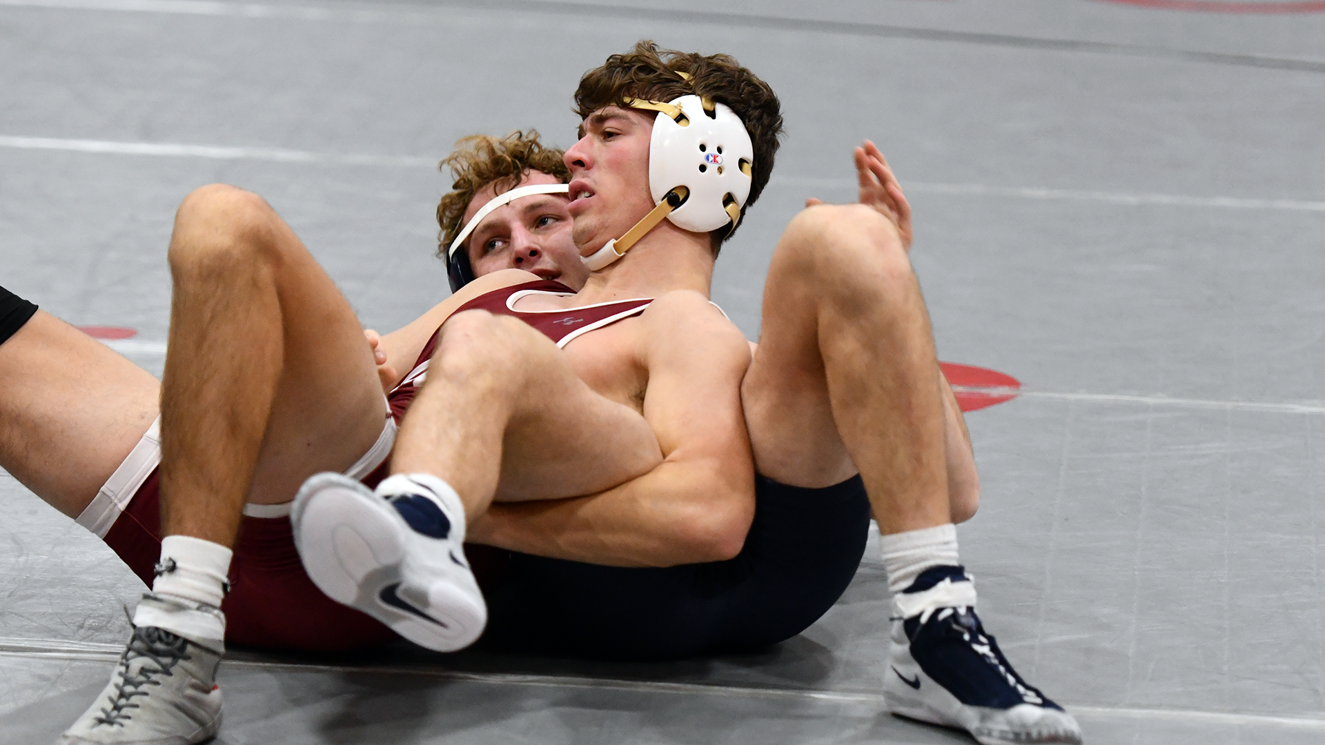 A wrestler in a red singlet and white headgear leans back on his opponent