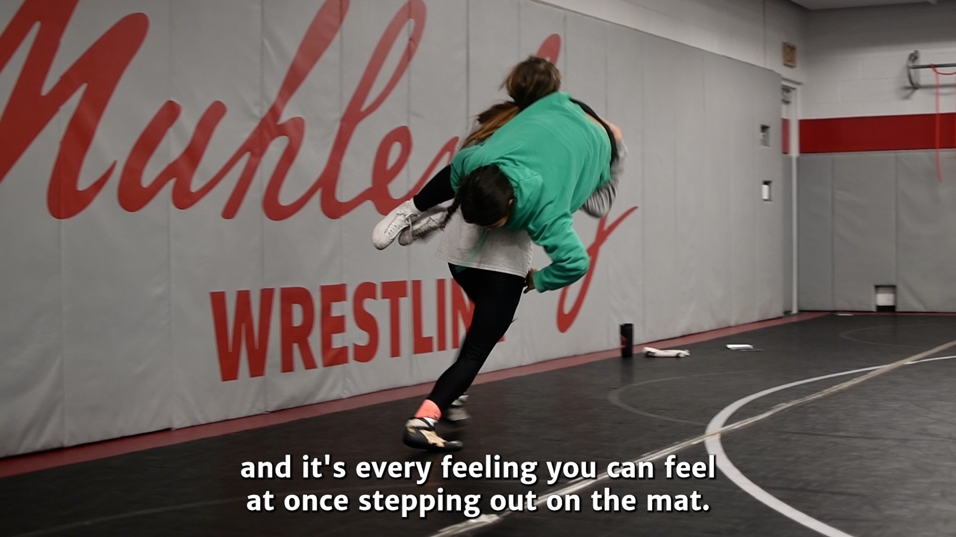 Two women's wrestlers practice in a room with Muhlenberg wrestling written on the wall and the text 