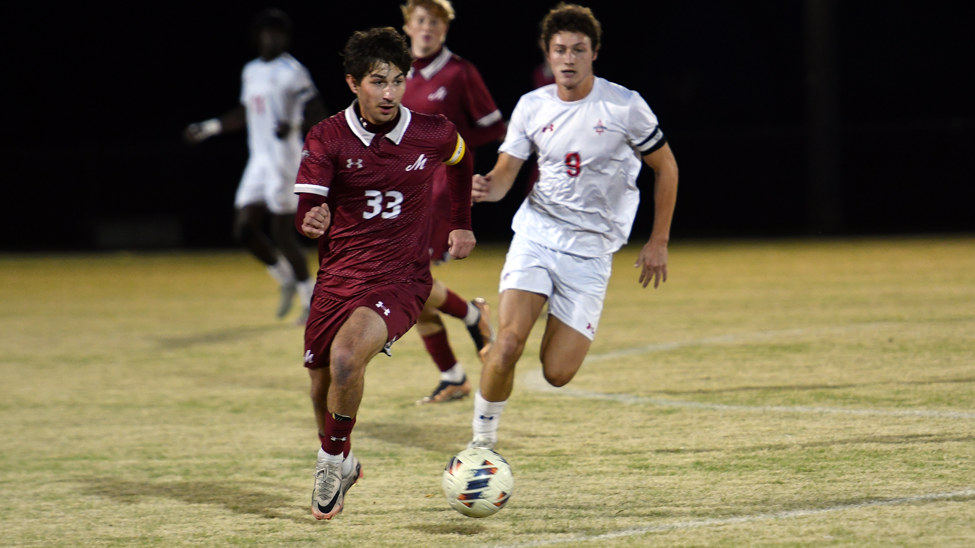 A soccer player in a red jersey dribbles while being trailed by a player in a white jersey