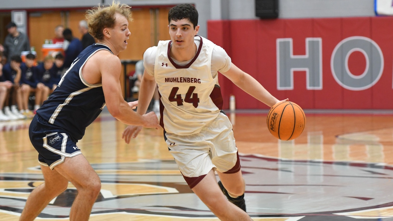 A basketball player in a white uniform drives past a defender in a blue uniform