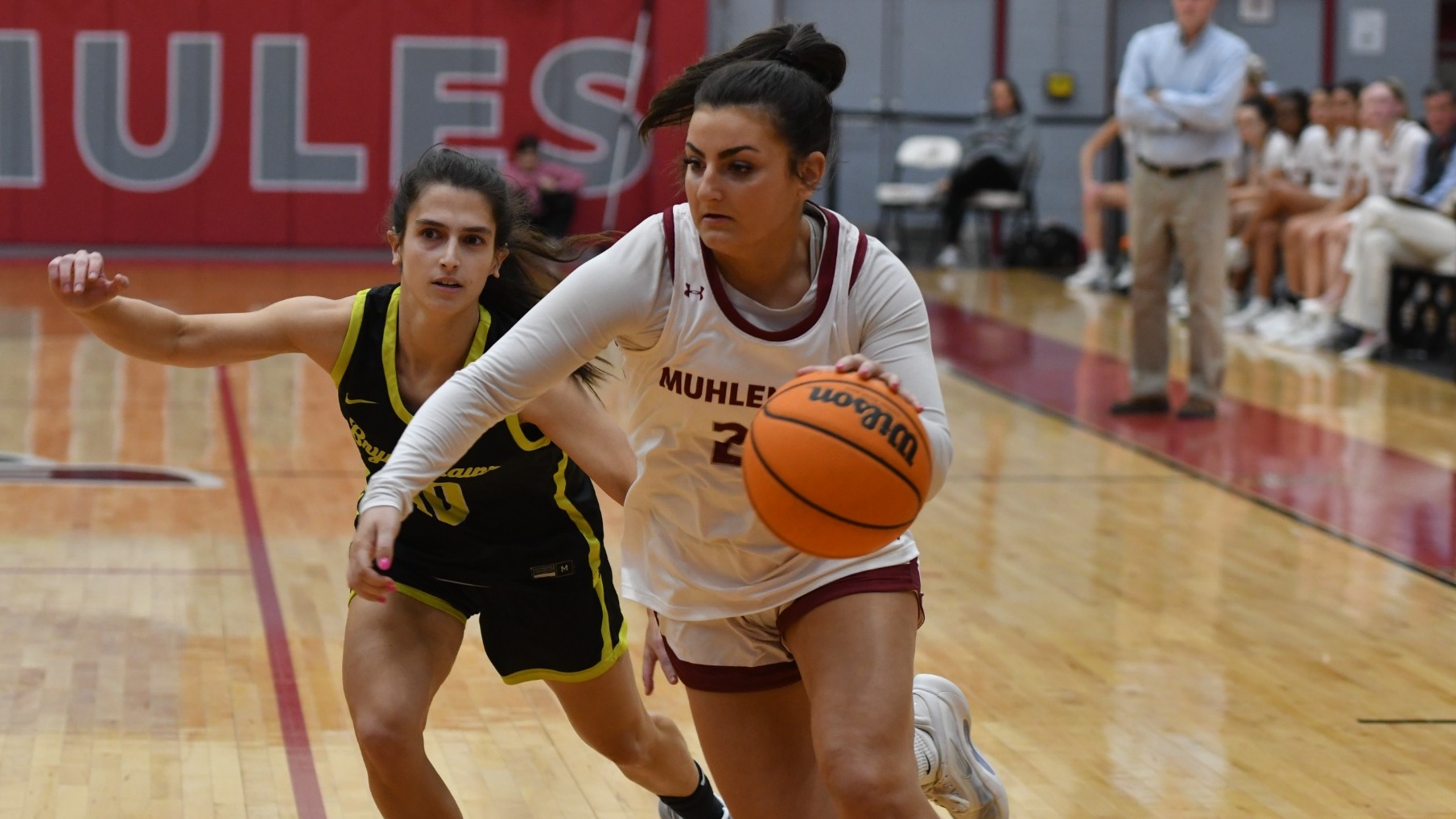 A basketball player drives to the basket in a white uniform while being guarded by a basketball player in a black uniform