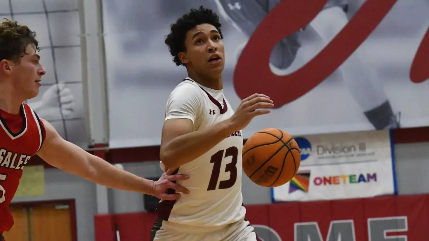 A basketball player in white with the ball drives to the basket while guarded by a player in red.