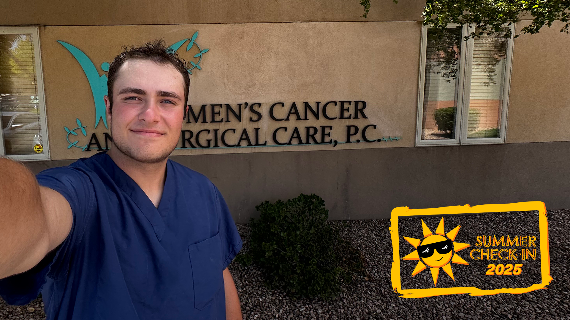 A young man in blue scrubs takes a selfie in front of a building with Women's Cancer and Surgical Care, P.C. on the side.