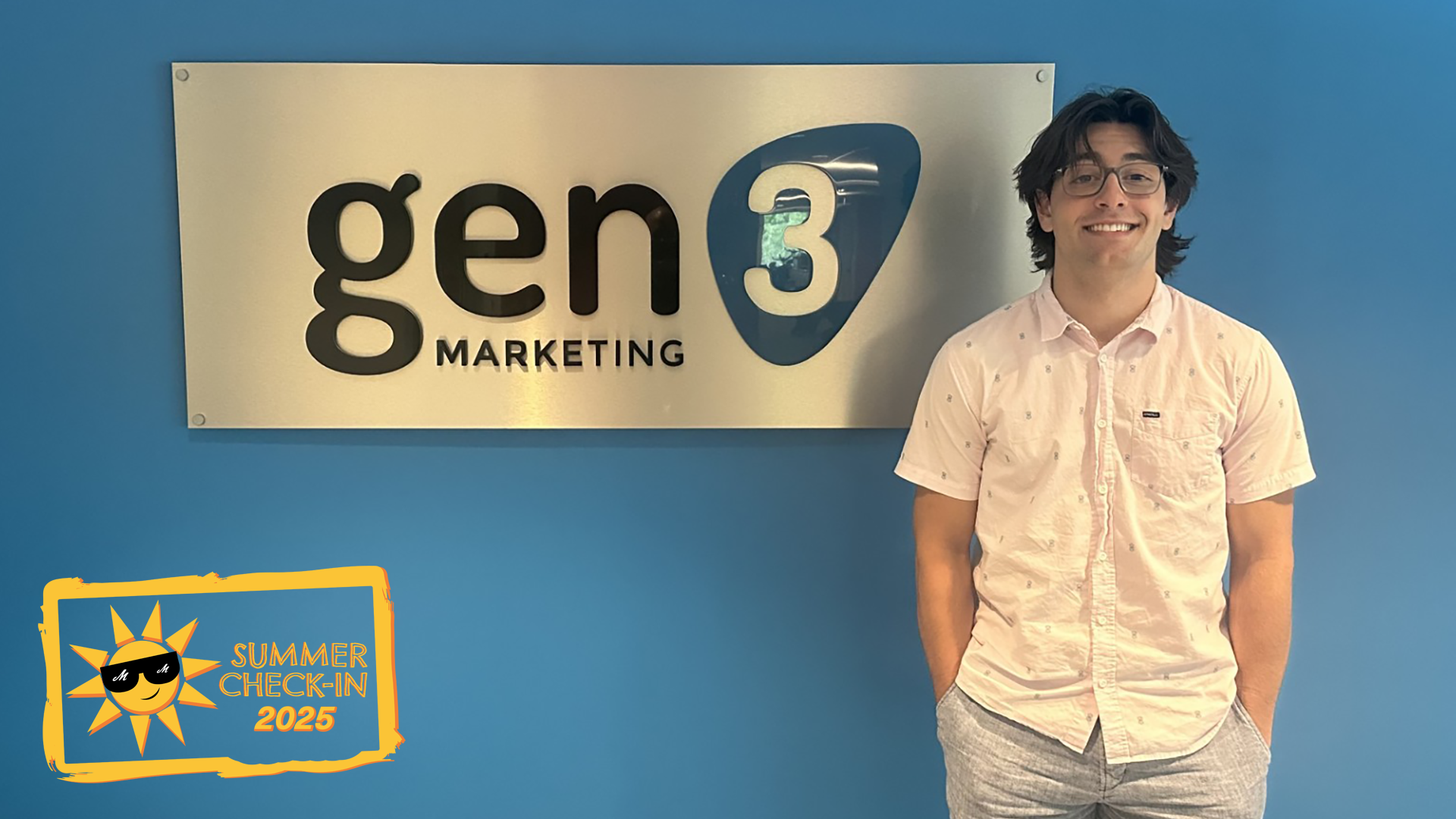 A young man in glasses and casual dress clothes stands in front of a blue wall with a sign reading gen 3 Marketing