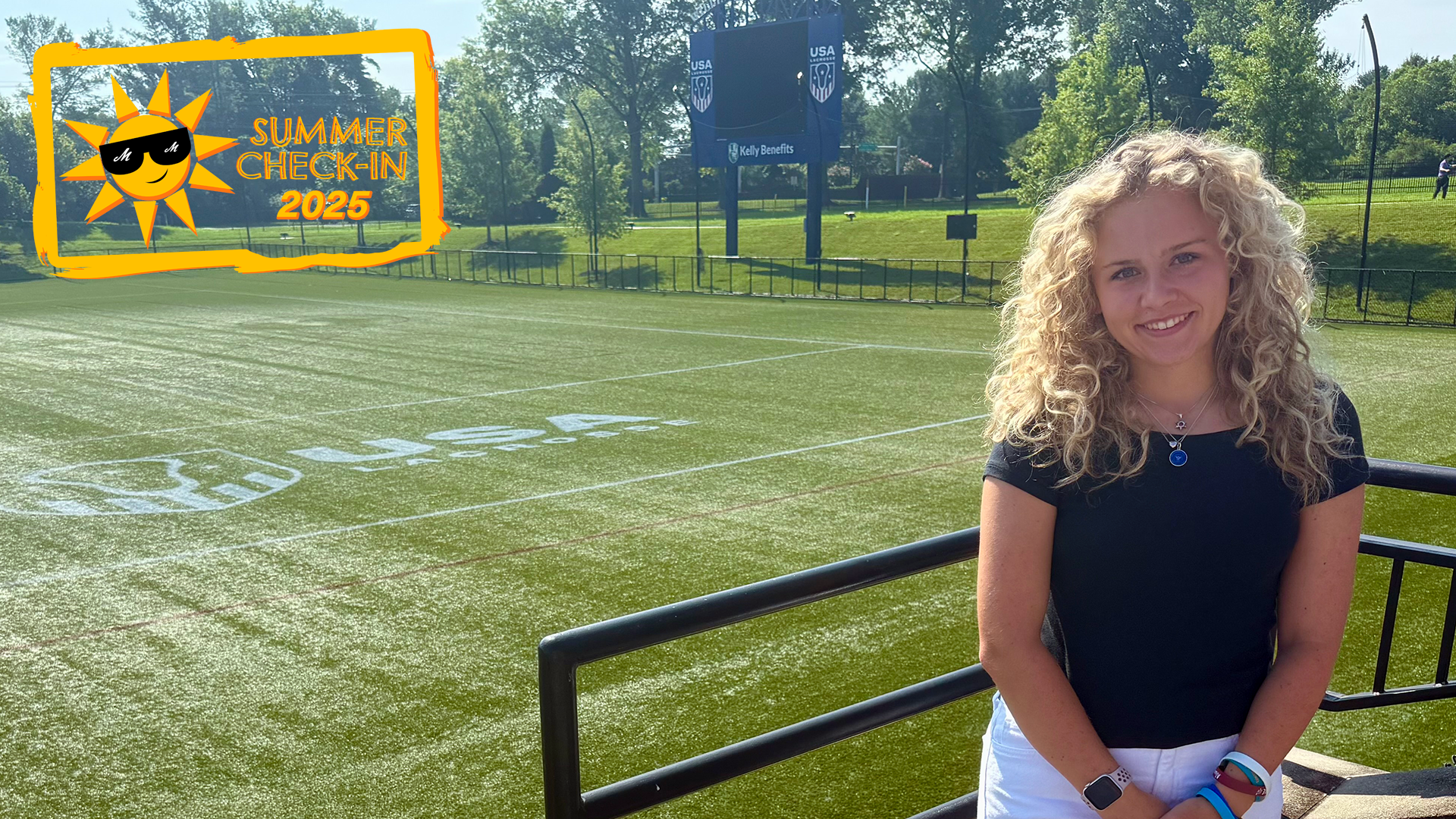 A young woman poses in front of a field and scoreboard with the USA Lacrosse logo on them