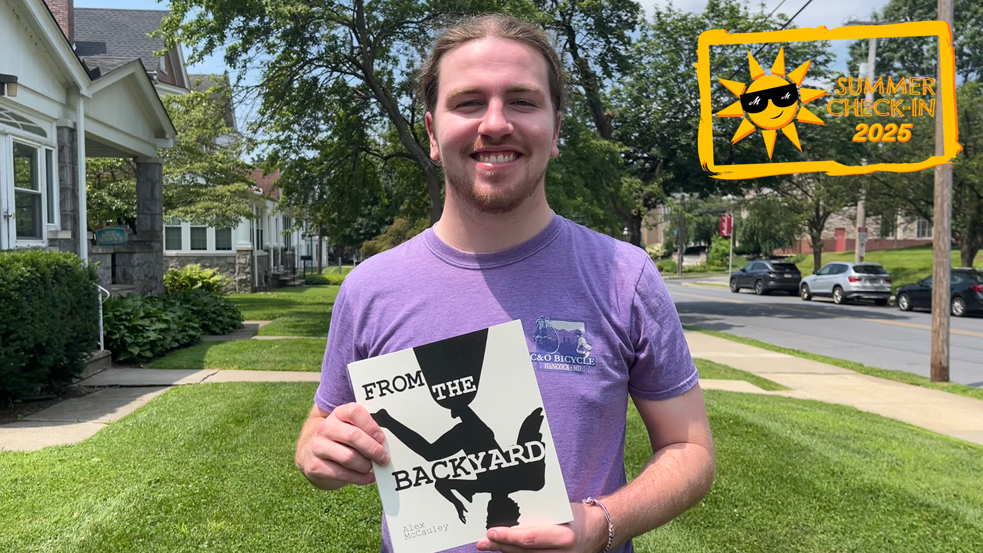 A young man in a purple t-shirt poses outdoors holding a book with the title From The Backyard