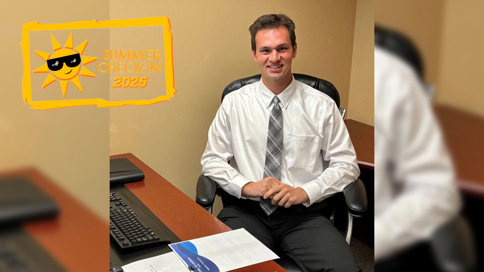 A young man in a white shirt and grey tie sits at an office desk