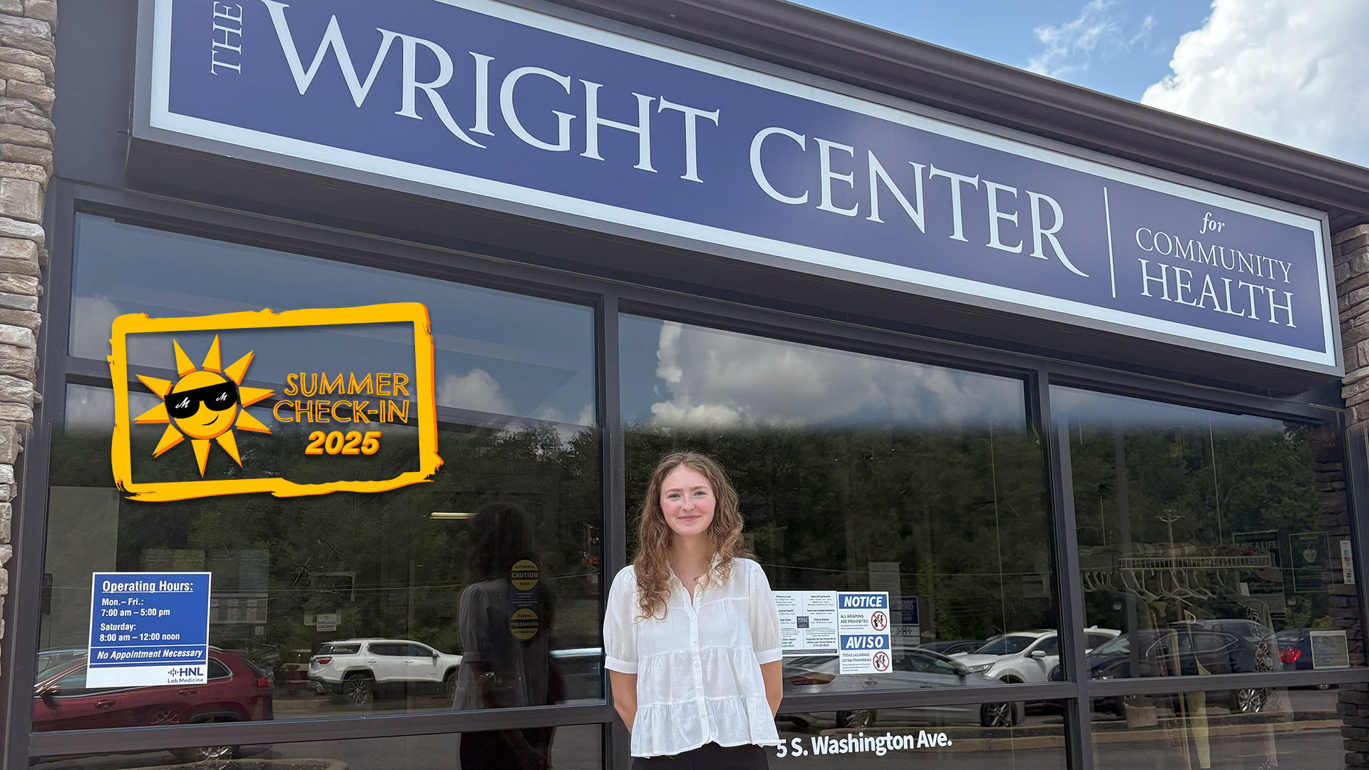 A young woman in a white shirt stands outdoor in front of a glass wall. A blue sign with The Wright Center for Community Health is at the top of the wall