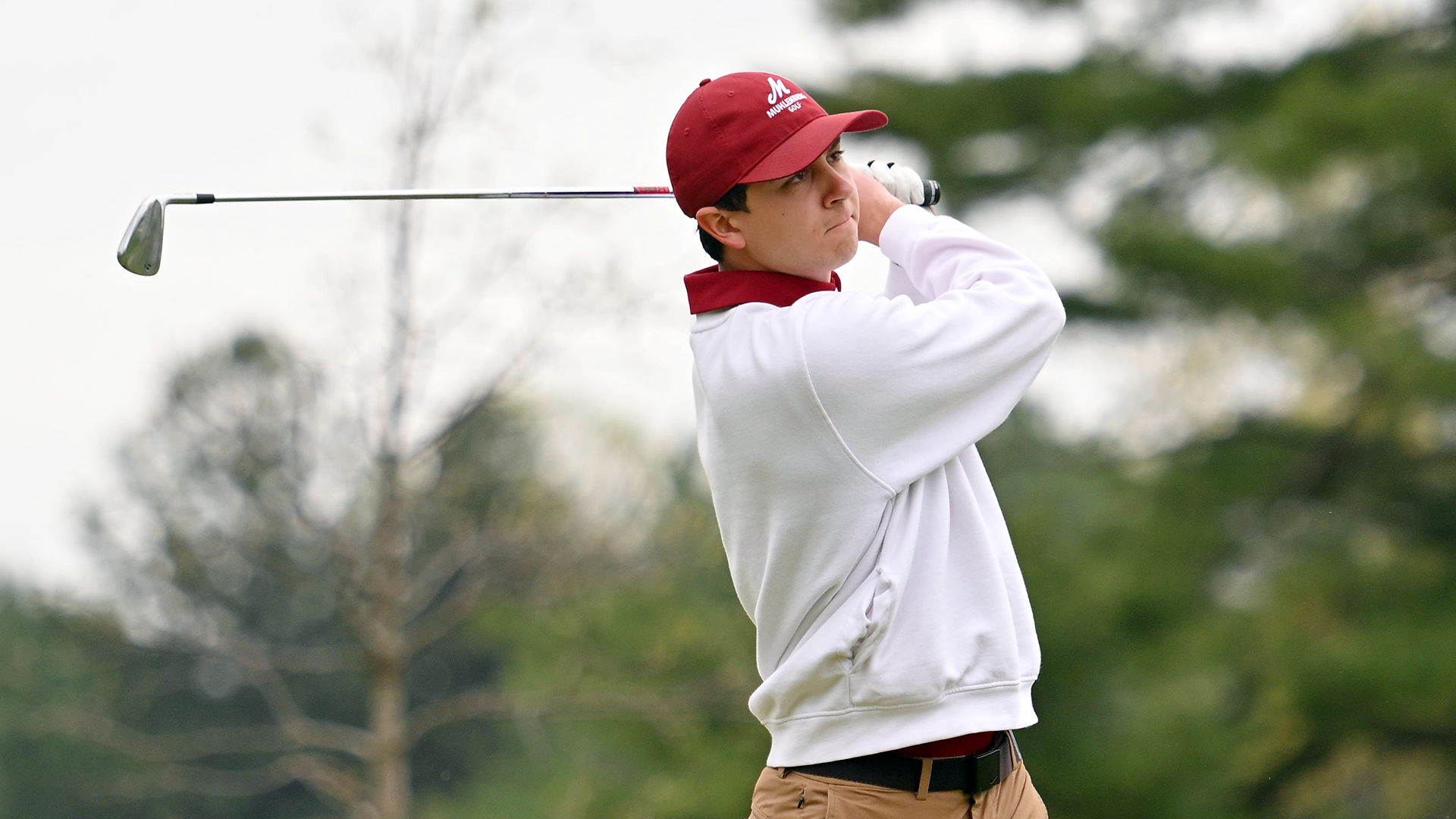 A golf player in a white sweatshirt and red hat follows through on his swing