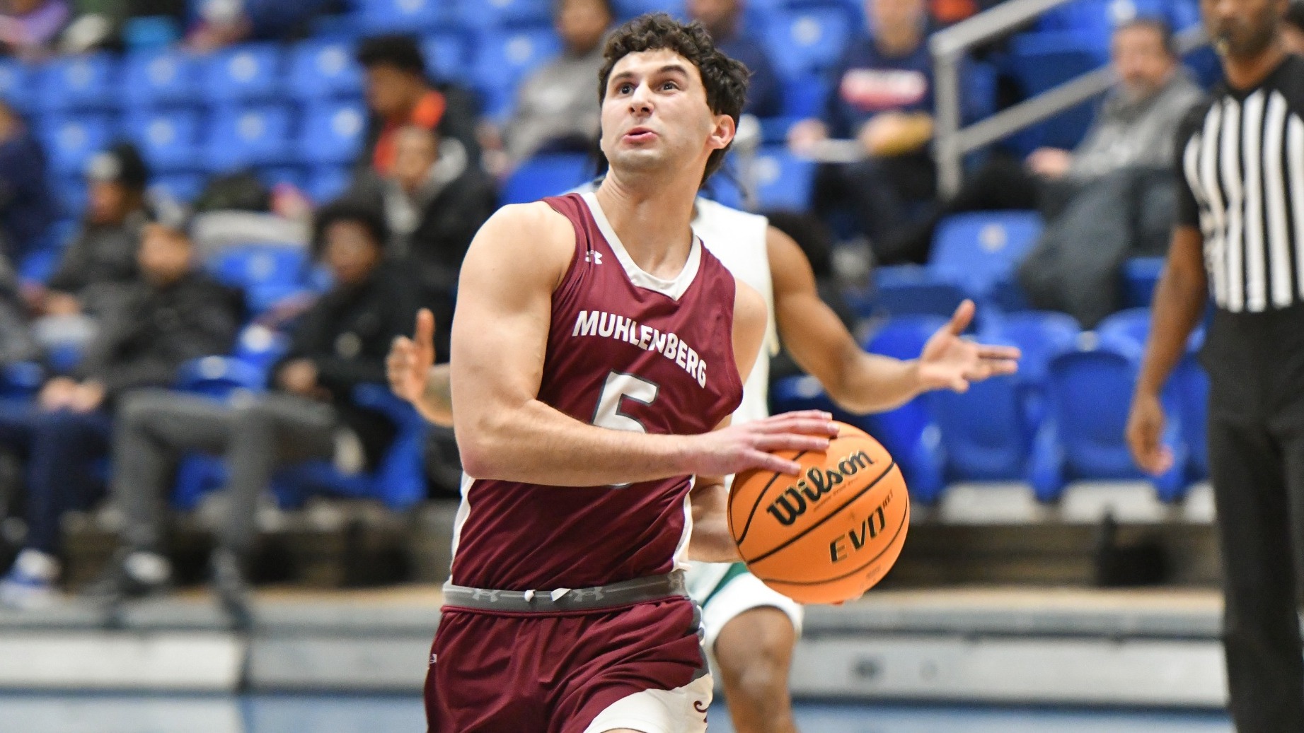 A basketball player in a maroon Muhlenberg jersey drives toward the basket while holding the ball with blue bleachers visible in the background.