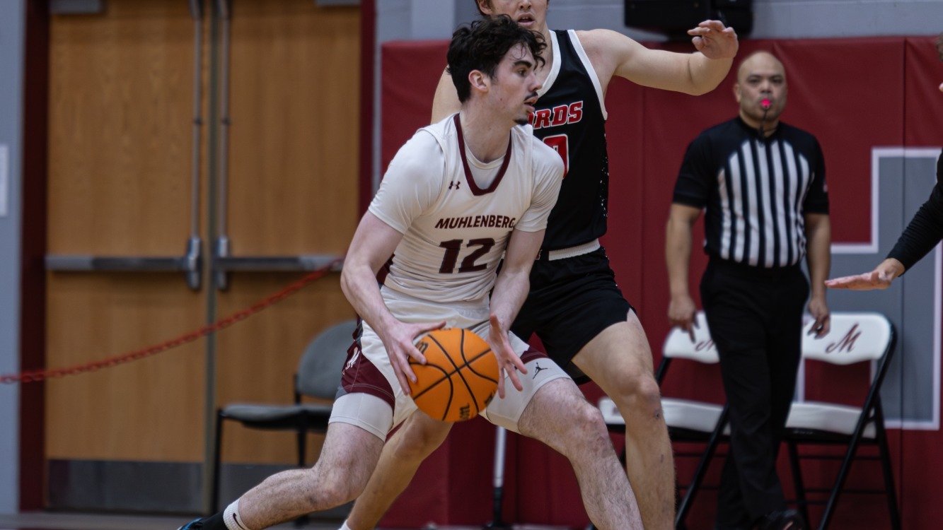 A Muhlenberg men’s basketball player wearing white protects the ball while driving toward the basket as a defender pressures him.