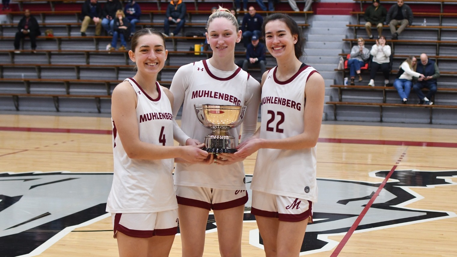 Three women's basketball players in white uniforms pose with a silver bowl trophy 