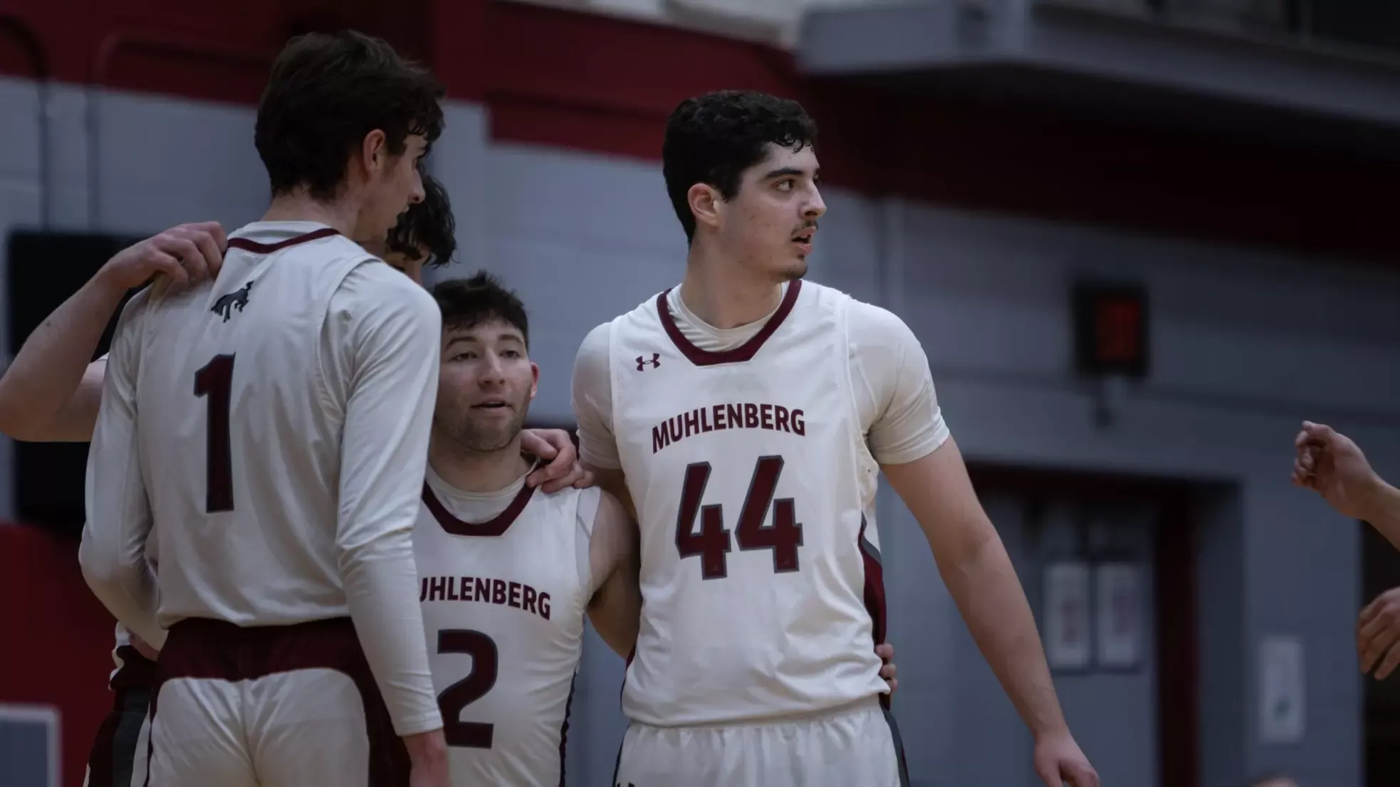 Three Muhlenberg basketball players wearing white jerseys stand together in a brief huddle on the court during a game.