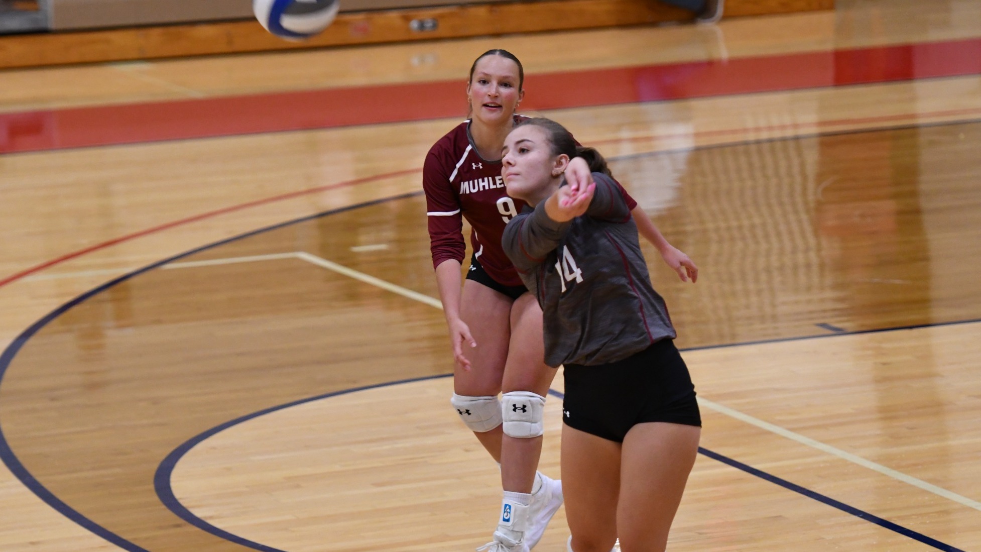 A Muhlenberg College volleyball player in a gray jersey dives forward to receive a volleyball as a teammate in a maroon jersey watches from behind on the indoor court.