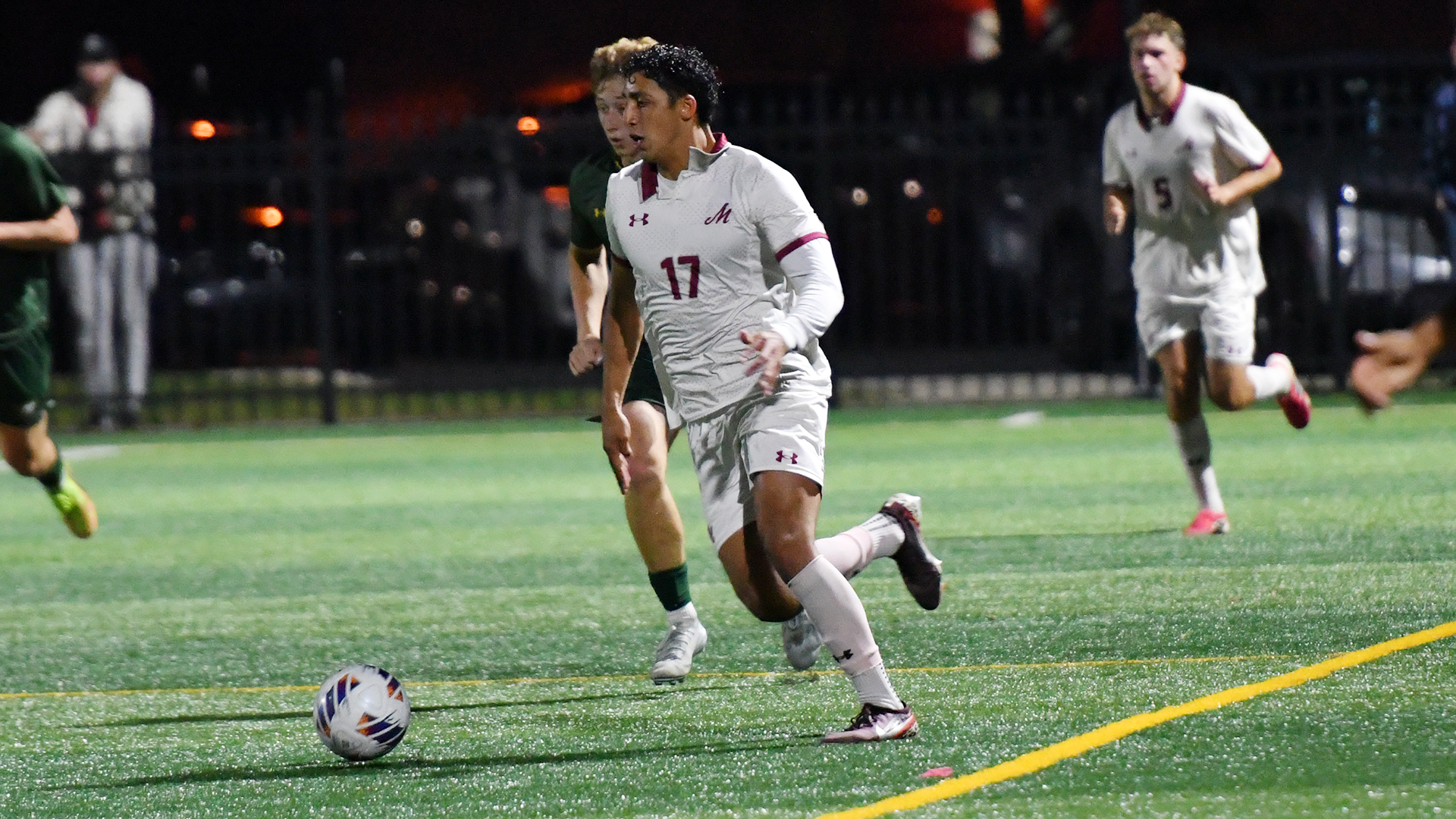 a soccer player in a white jersey dribbling past one in a green jersey in a night game on a turf field