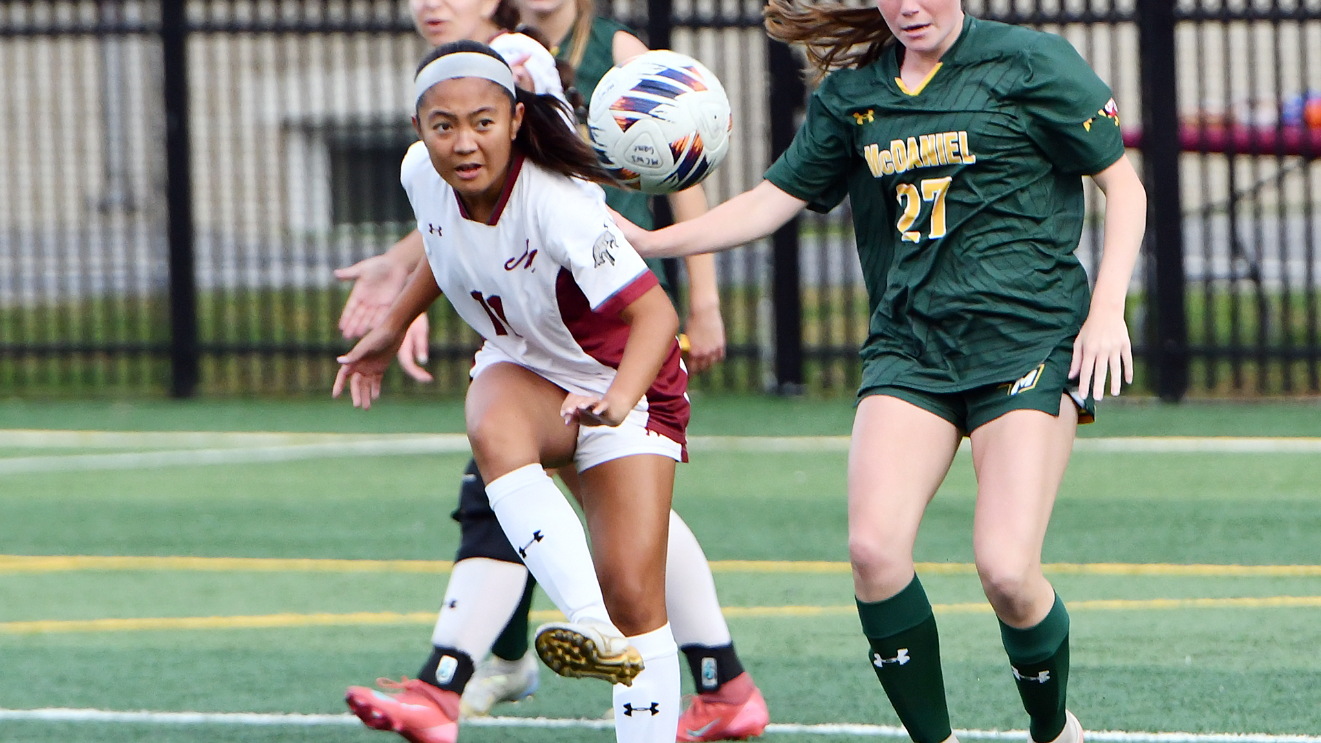 a soccer player in a white uniform kicking the ball while being guarded from behind by a player in a green jersey
