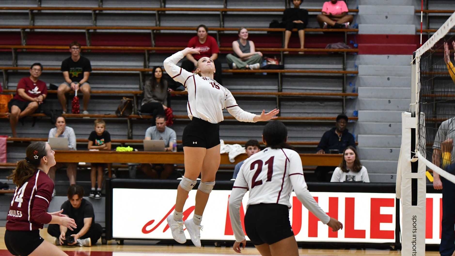 A volleyball player in a white uniform rises to spike the ball in a gym full of fans, with her teammates nearby