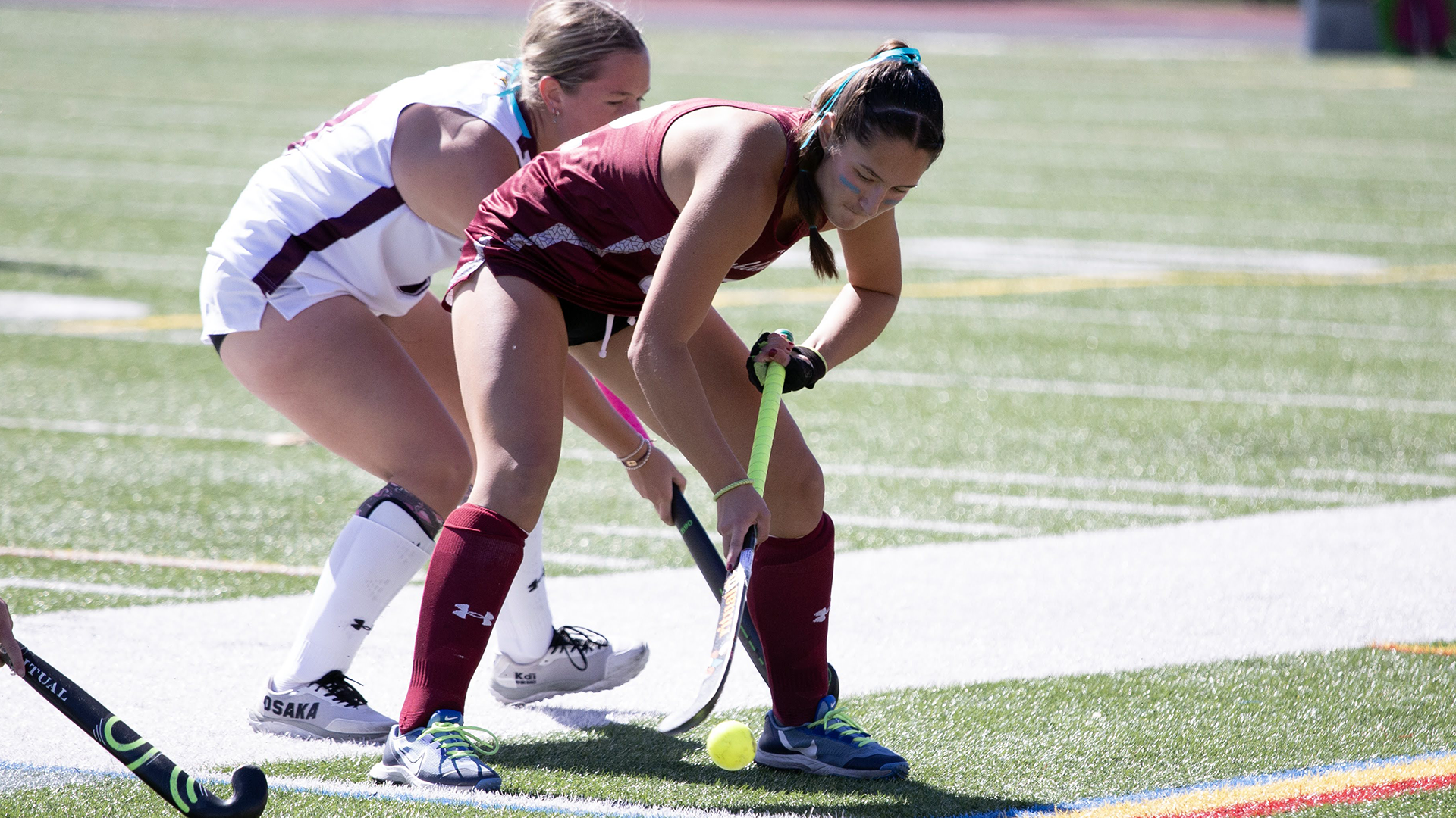 a field hockey player in a red jersey battling for the ball with a player behind her in a white jersey on a sunny day