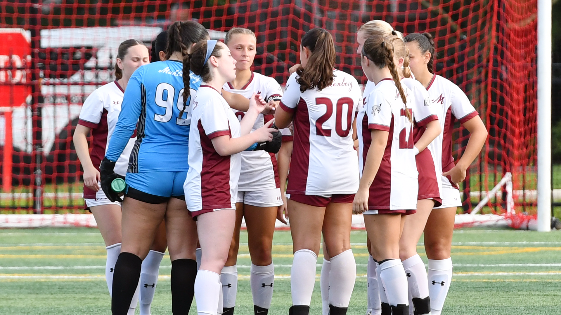women's soccer players in white uniforms standing in a huddle with the net behind them