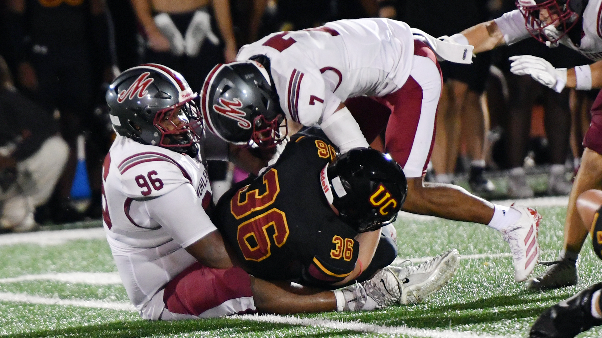 two football players in white uniforms and silver helmets combining to tackle a player in a black jersey and black helmet at night on a turf field