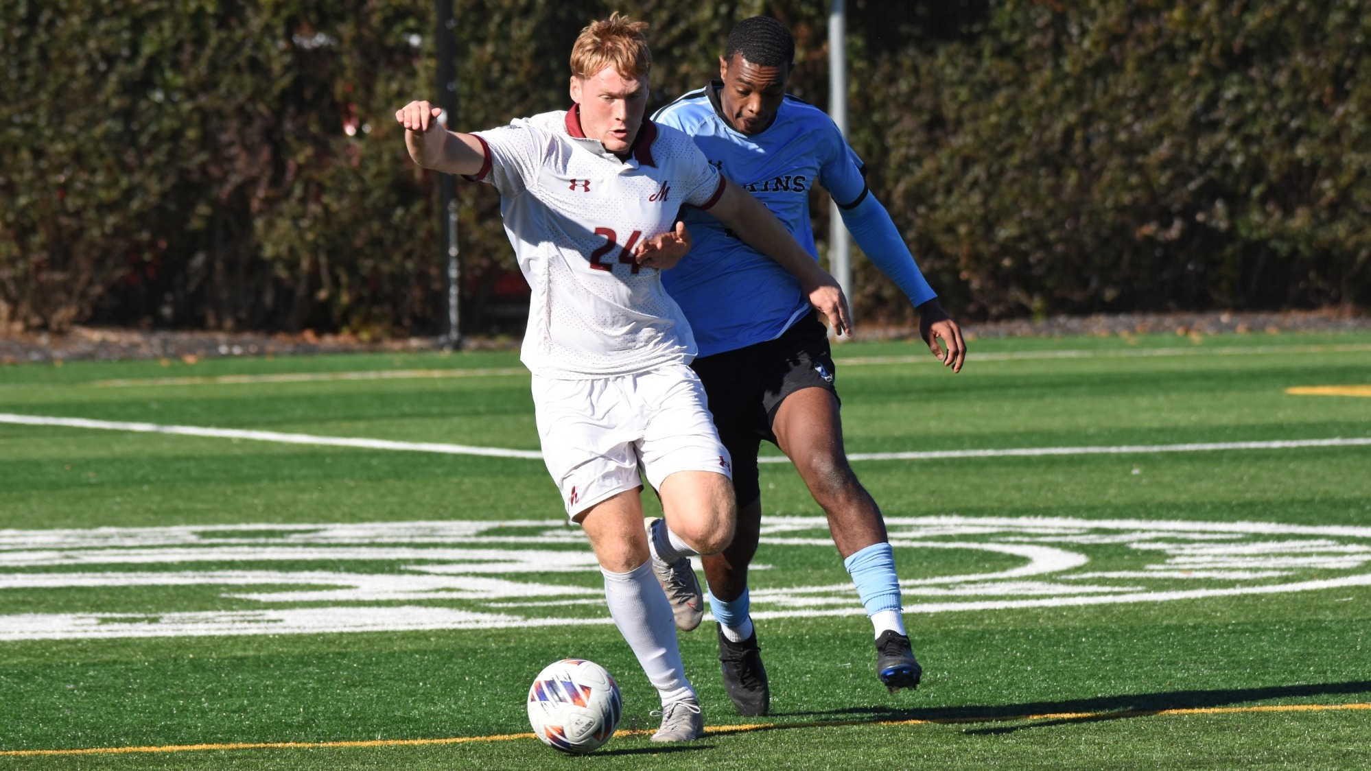 A soccer player in a white uniform battles for the ball with a soccer player in a blue uniform right behind him. 