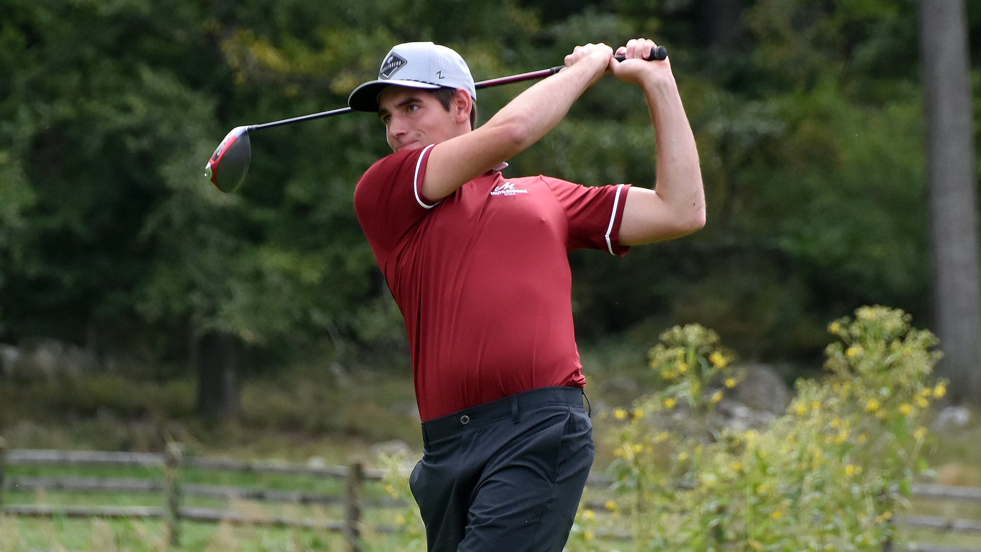 a golfer in a red shirt, black slacks, and a grey hat following through on a drive