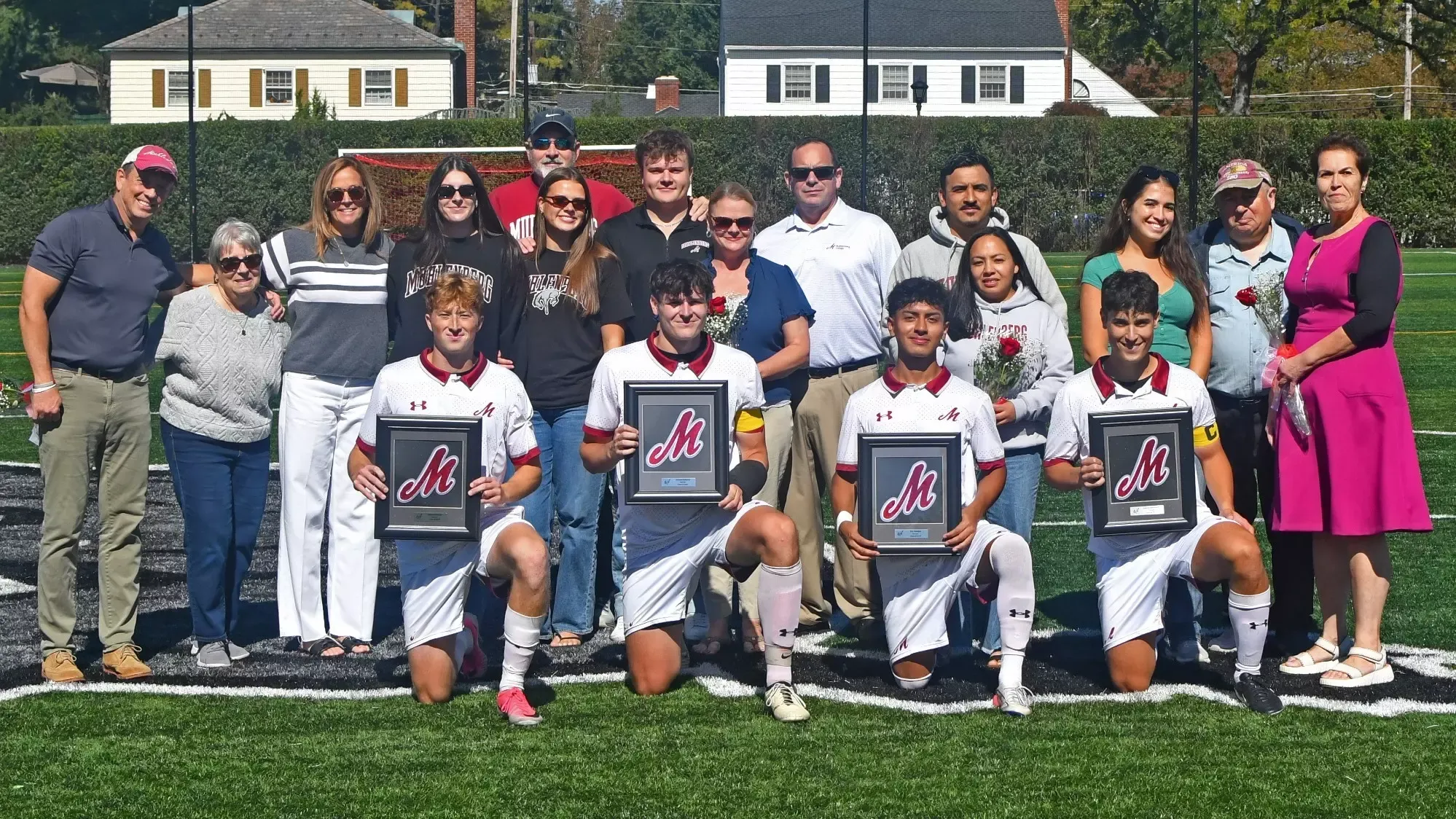 A group of four Muhlenberg soccer players kneel on a field holding framed awards, surrounded by family and supporters.