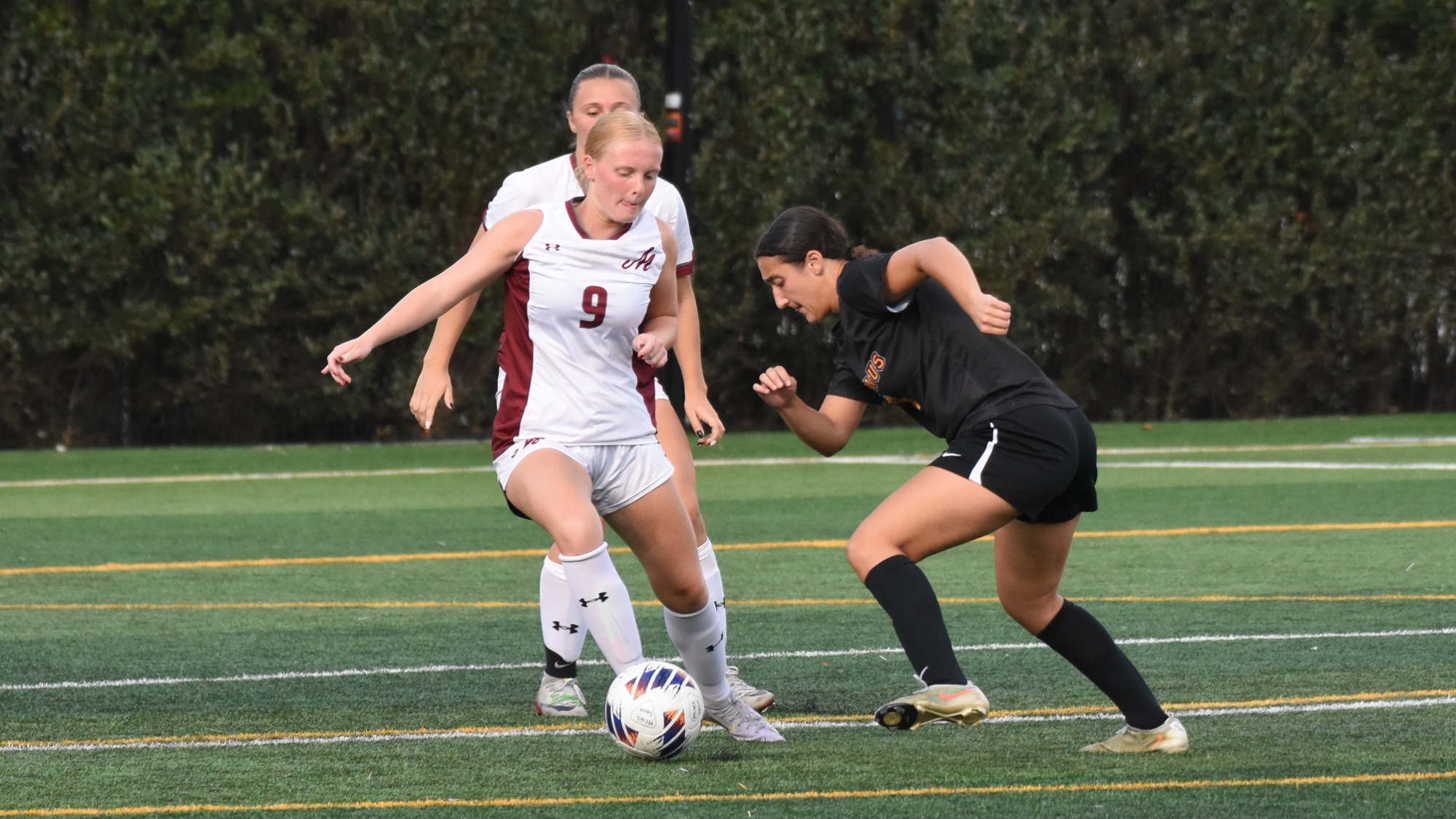 A Muhlenberg women’s soccer player in white battles an opposing player in black for possession of the ball on the field.