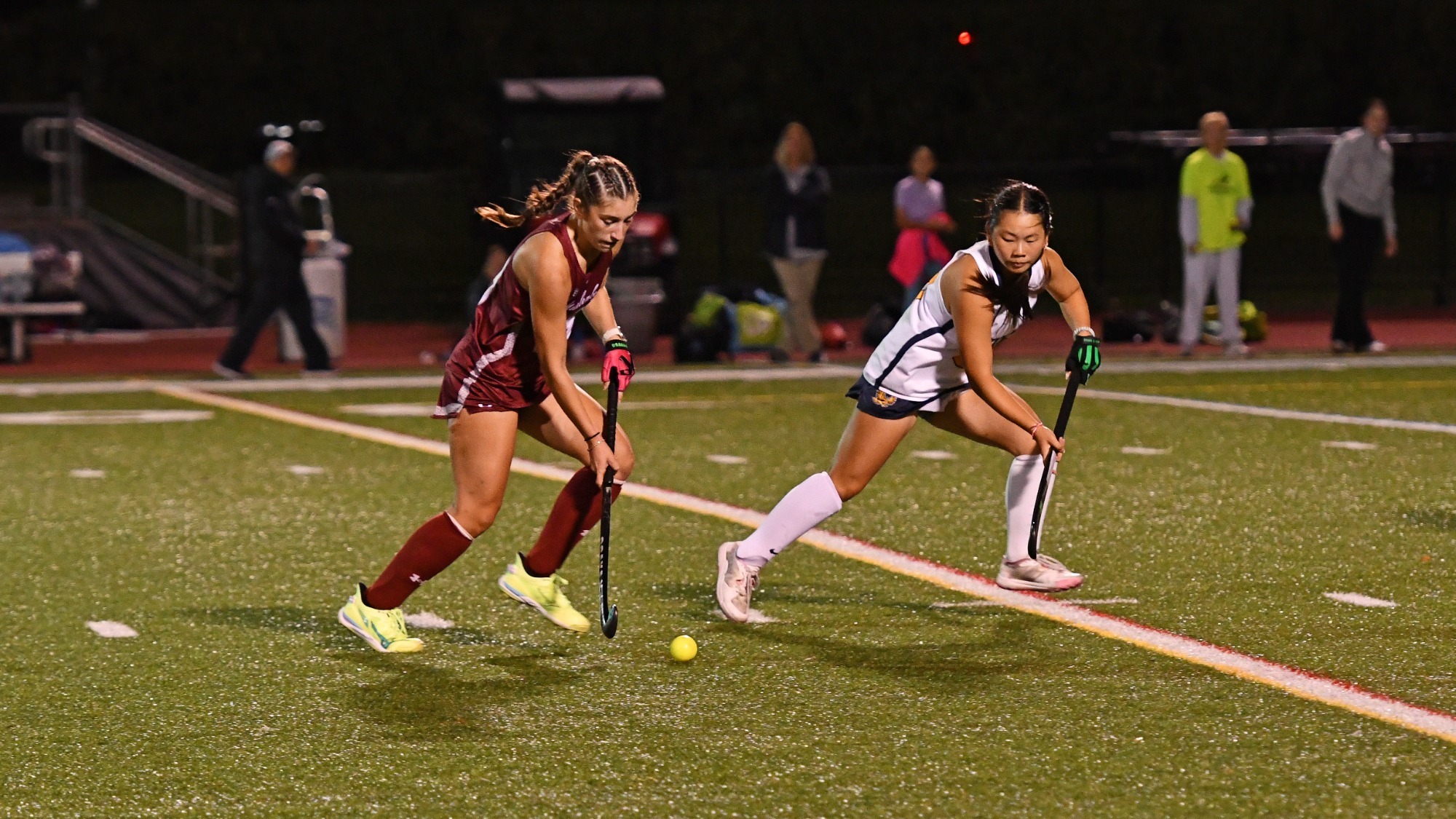 A field hockey player in red dribbles the ball up the field while a player in white guards her. 