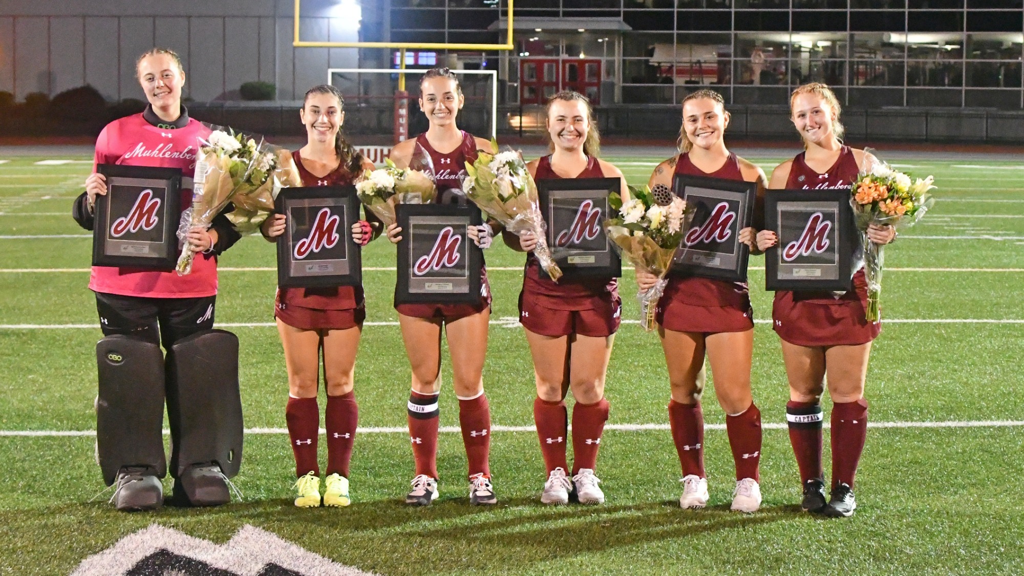 Six Muhlenberg College field hockey players pose on the turf at night with framed Muhlenberg logos and bouquets of flowers during a senior recognition celebration.