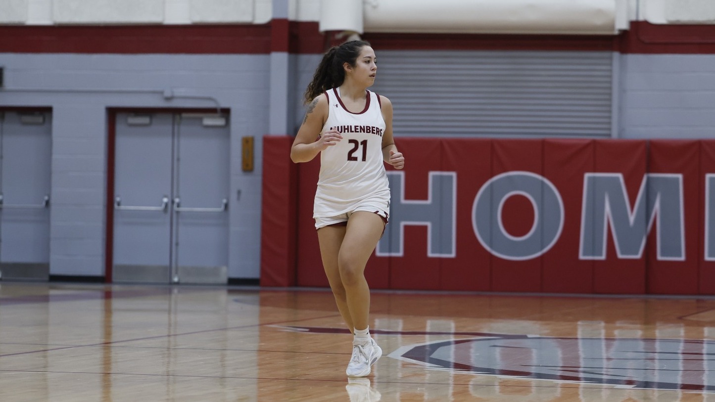 A Muhlenberg women’s basketball player in a white jersey runs up the court in a gym with red wall padding.