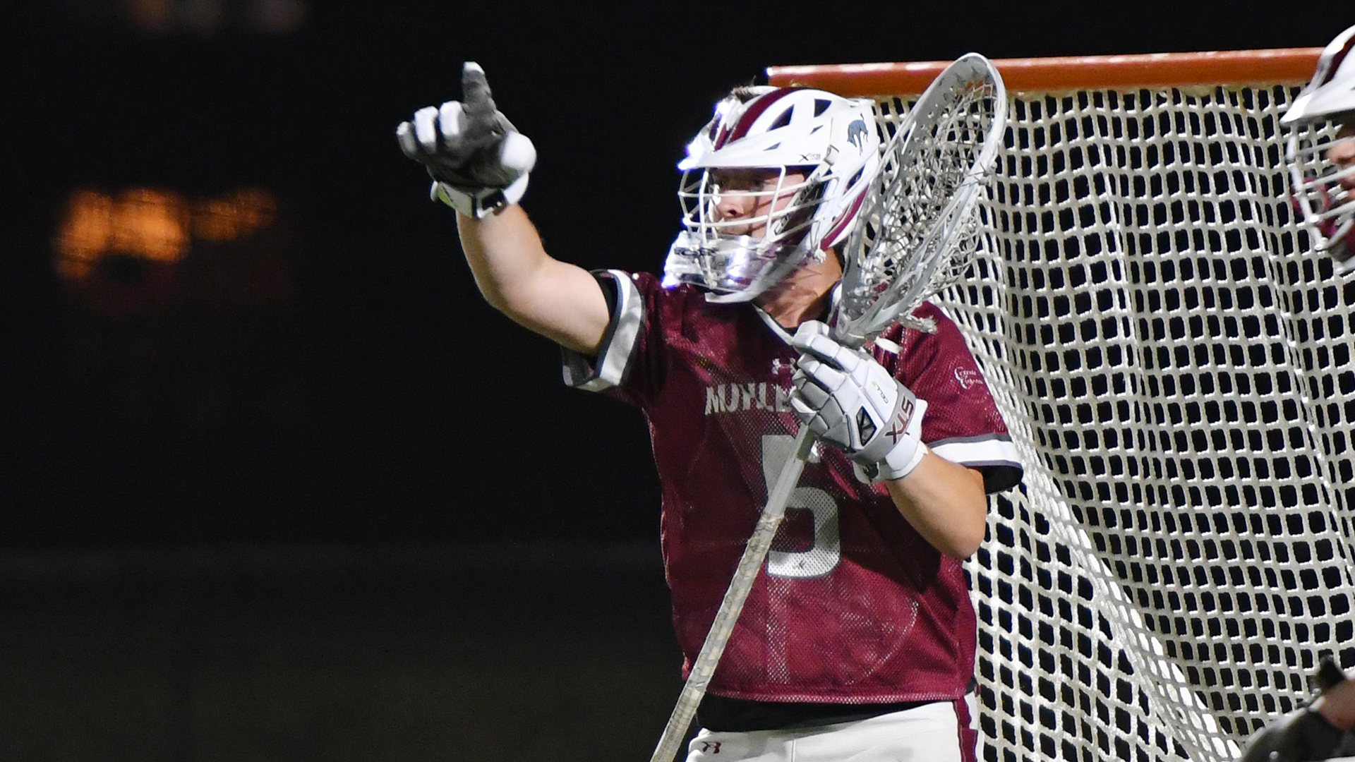 a men's lacrosse goalie with a red jersey points with his right hand while standing in goal