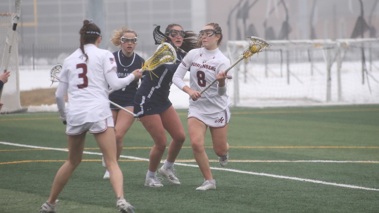 A Muhlenberg women’s lacrosse player in a white jersey carries the ball while weaving between two defenders in navy on a foggy turf field.