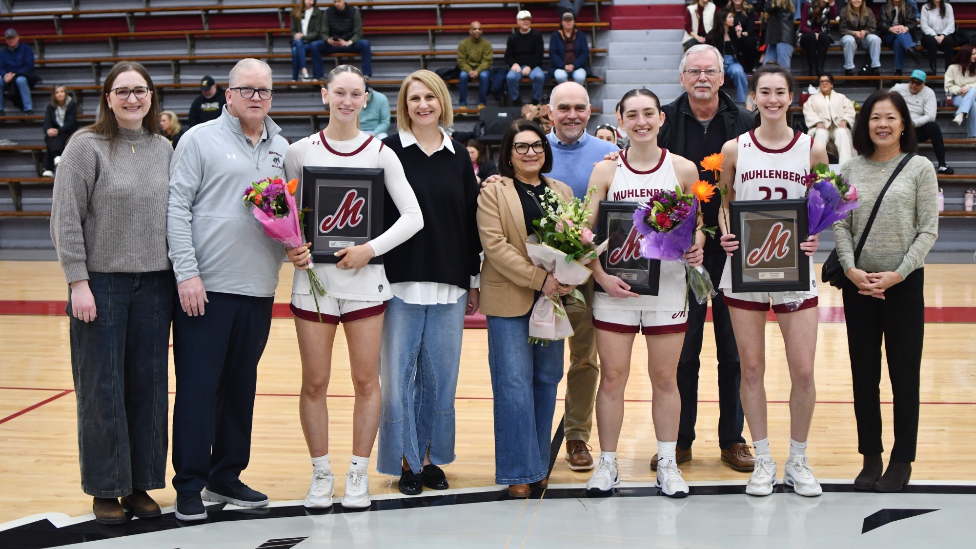 Basketball players and their families stand together on the court holding framed plaques during a senior night ceremony, with fans seated in the bleachers behind them.