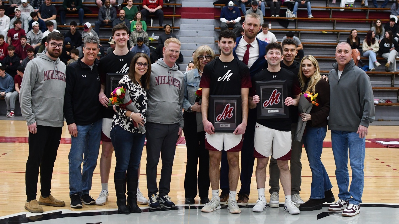 Basketball players and their families stand together on the court holding framed plaques during a senior night ceremony, with fans seated in the bleachers behind them.