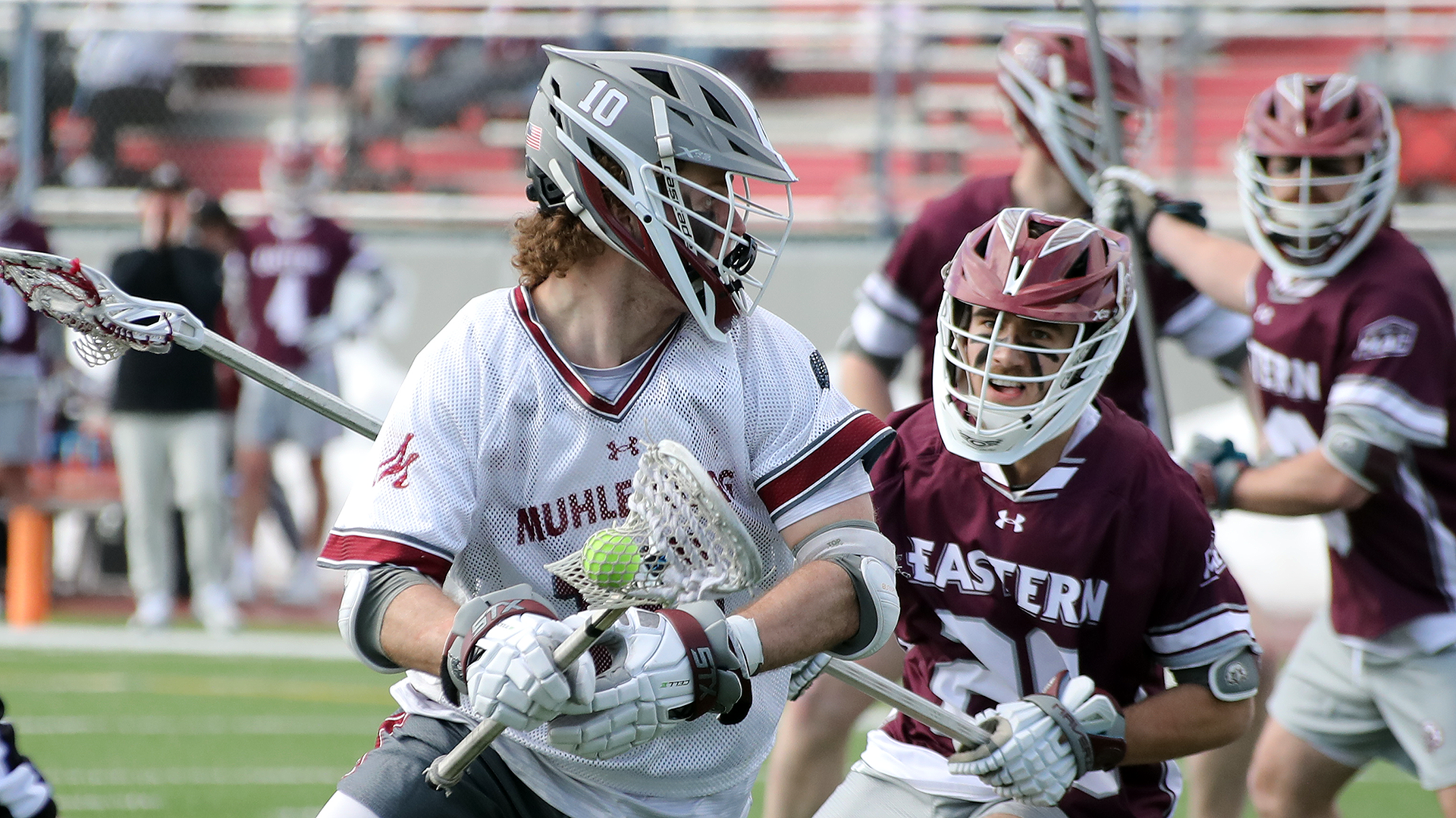 a men's lacrosse player in a white uniform and grey helmet guarded by a player in a red uniform and red helmet