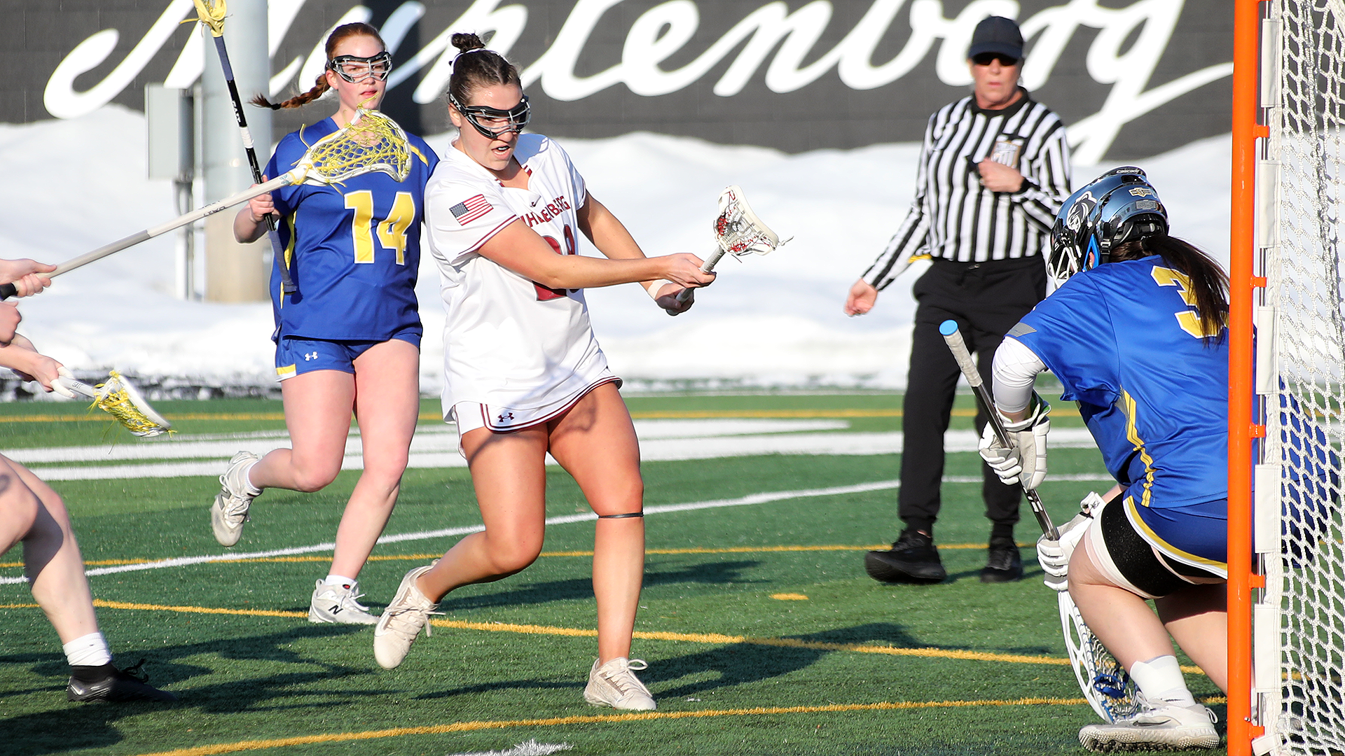 a women's lacrosse player in a white jersey shooting the ball at a net guarded by a goalie in a blue jersey and blue helmet