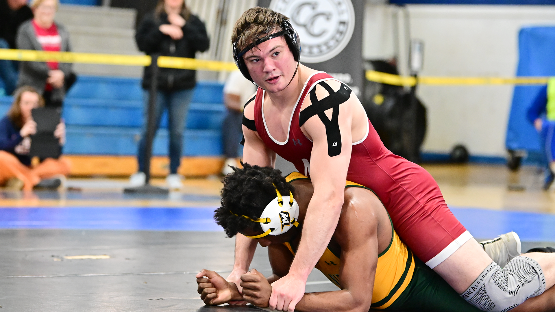 a wrestler in a red singlet looks up while riding a wrestler in a gold and green singlet