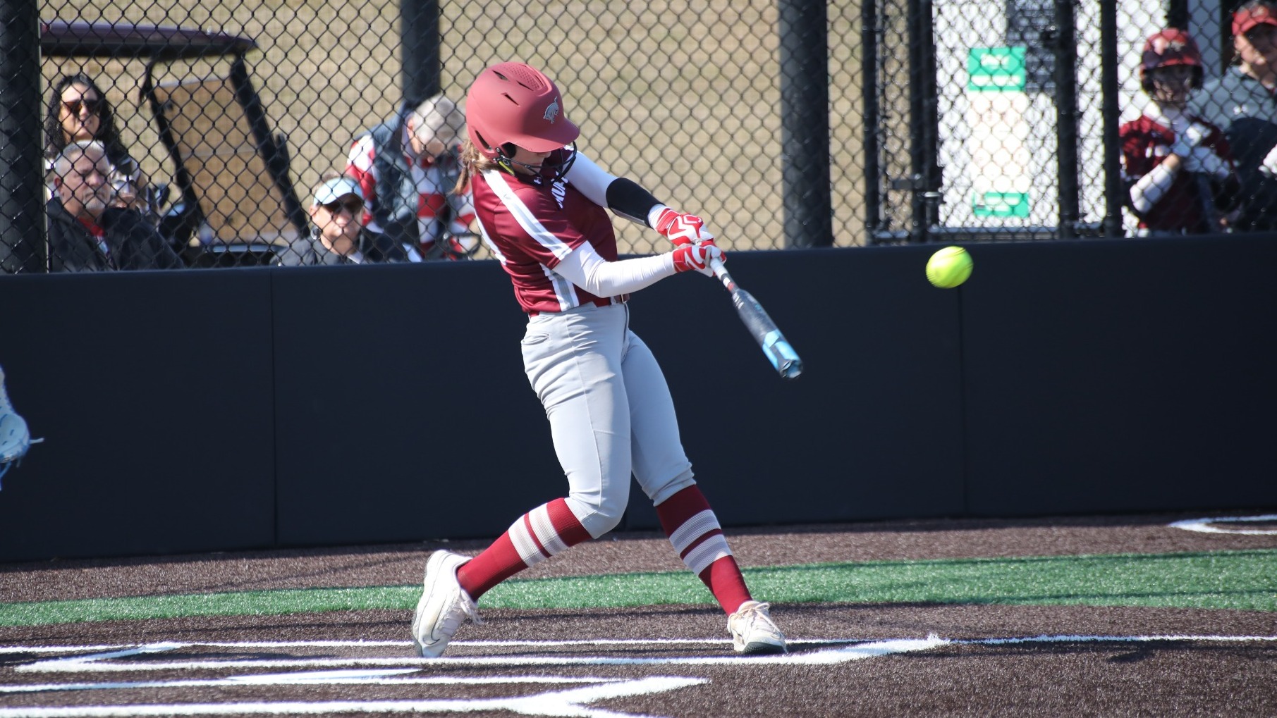 A softball player in a maroon helmet and jersey swings and hits the softball.