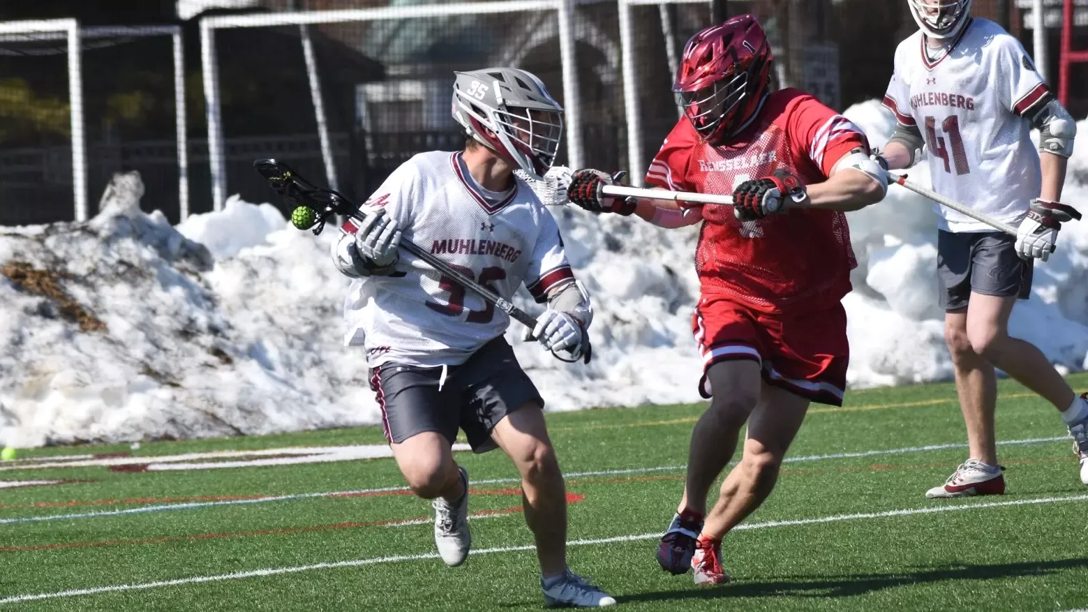 A Muhlenberg men’s lacrosse player in a white jersey dodges a Rensselaer defender in red during a game on a turf field with snow piled along the sidelines.