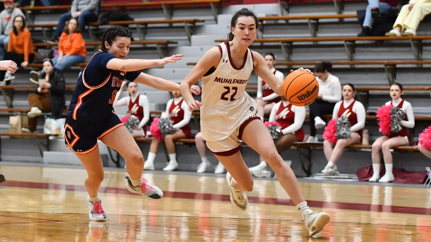A basketball player in a white Muhlenberg jersey dribbles past a defender wearing a blue jersey