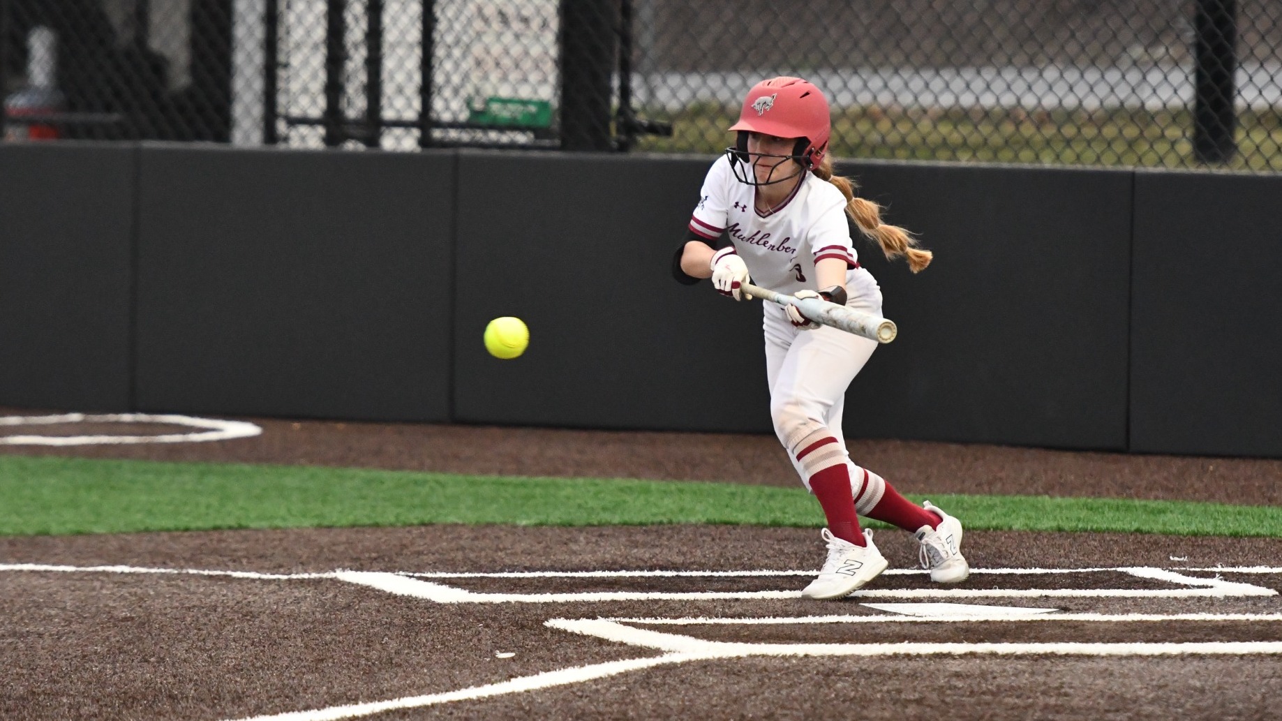 A softball player in a white uniform and red helmet squares to bunt as the ball approaches the bat at home plate.