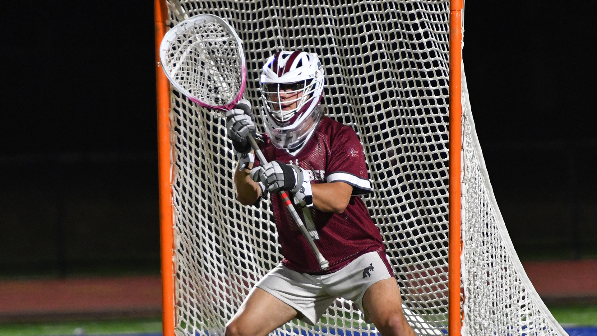 a lacrosse goalie in a red jersey and white shorts in his stance in front of the net