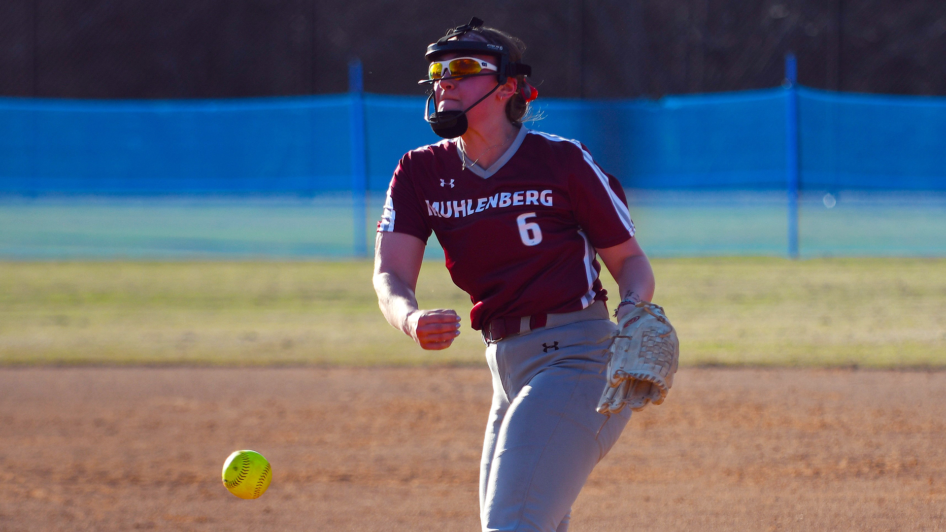 a softball pitcher in a red jersey and grey pants wearing number 6 releases a yellow softball
